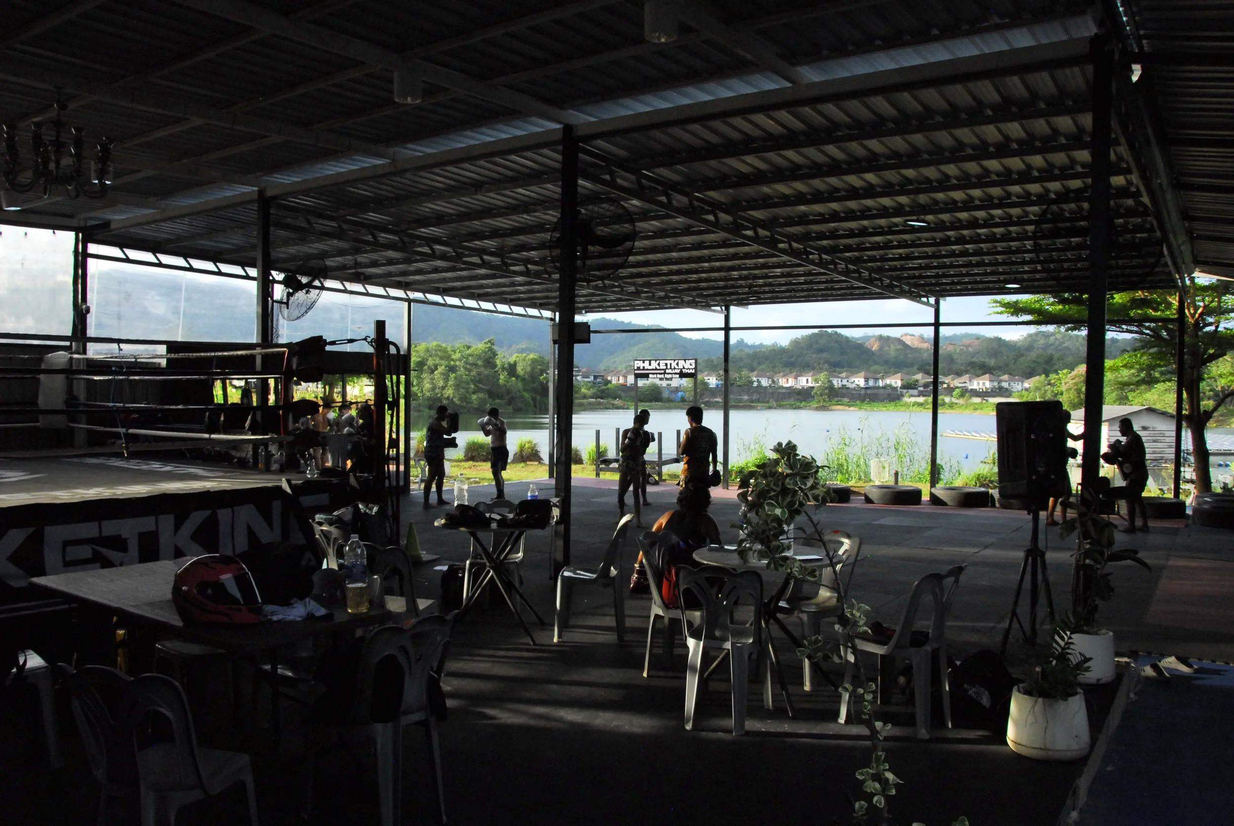 People training in a boxing gym with a boxing ring near a large open window overlooking a river and green hills.