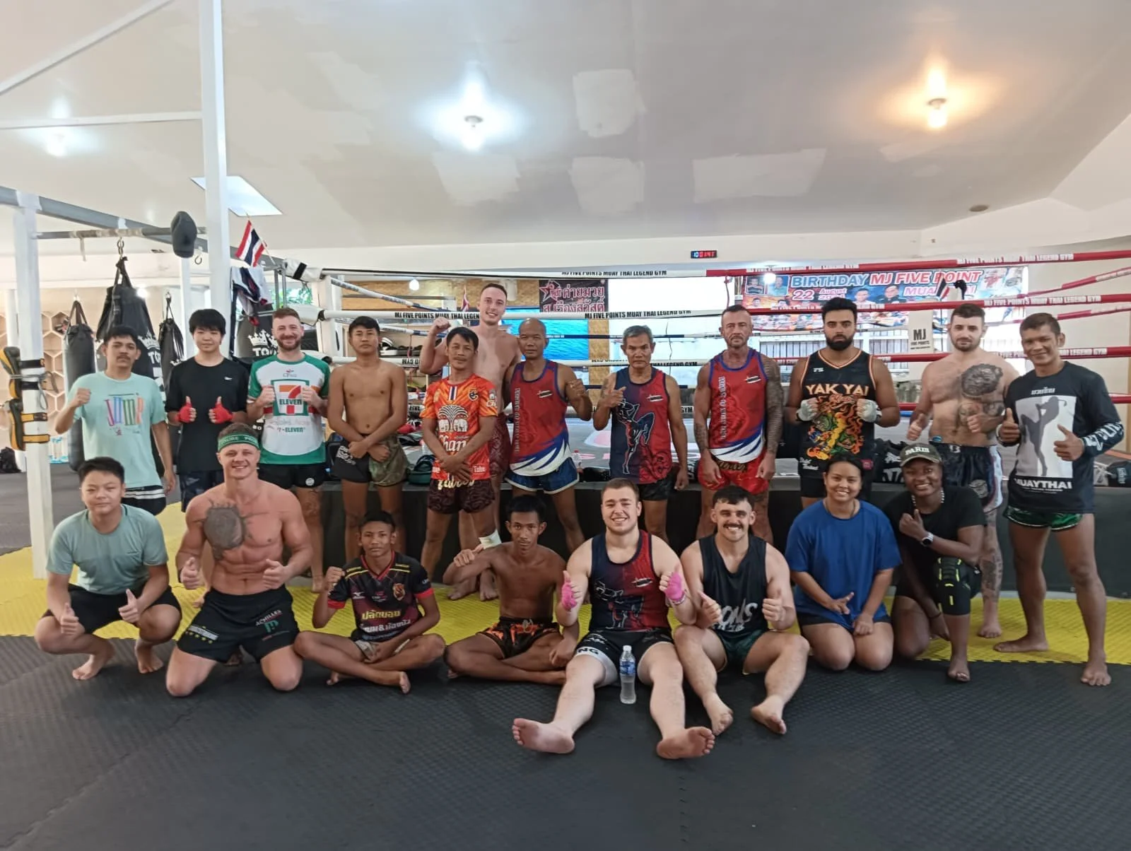 Group of Muay Thai fighters and trainers posing inside a boxing gym. They are wearing sports gear, shorts, and some with gloves. The background features a boxing ring and training equipment.