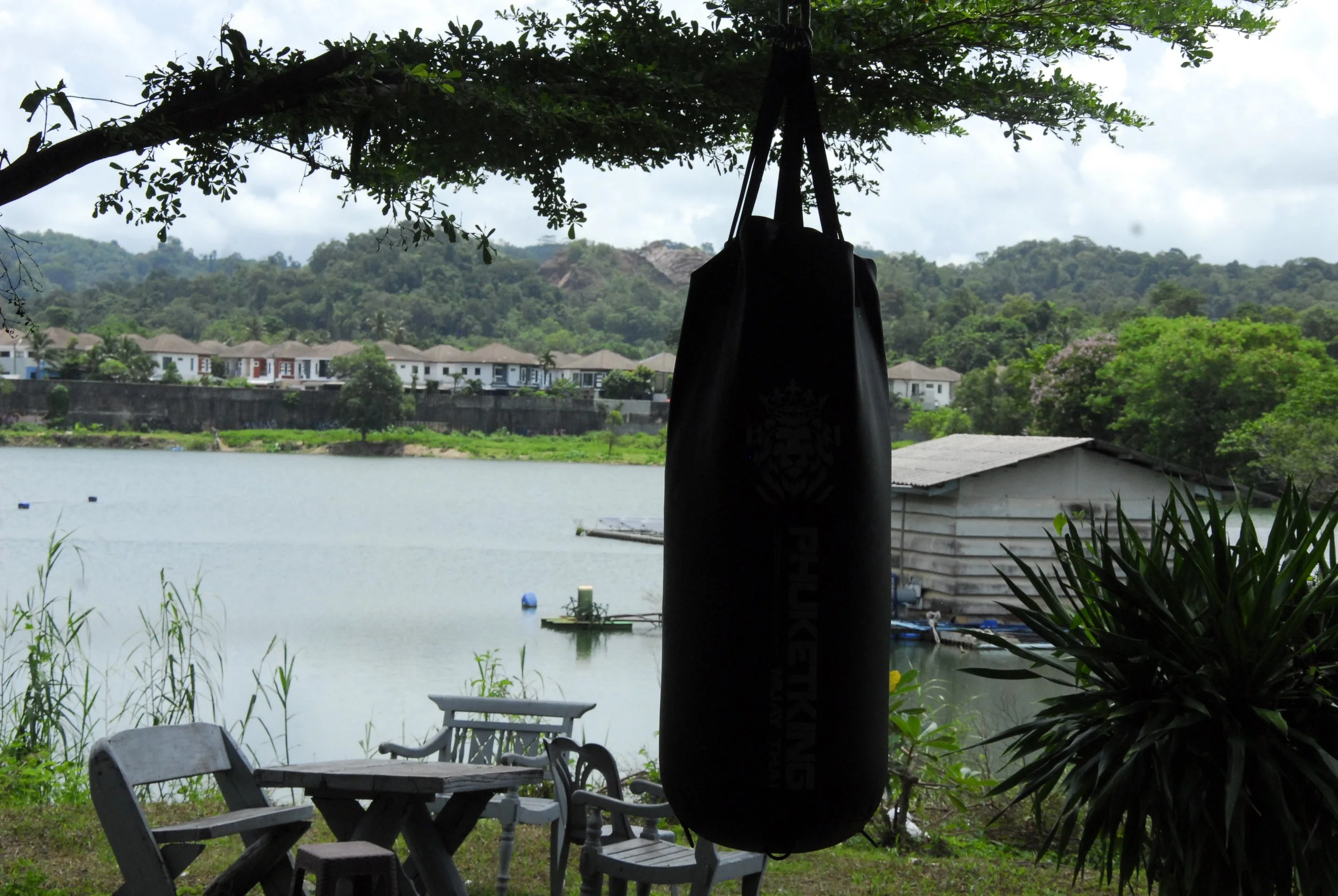 A lakeside scene with green trees, hills in the background, and residential buildings along the shore. In the foreground, there are two wooden chairs and a small table, with a hanging black punching bag partially blocking the view.
