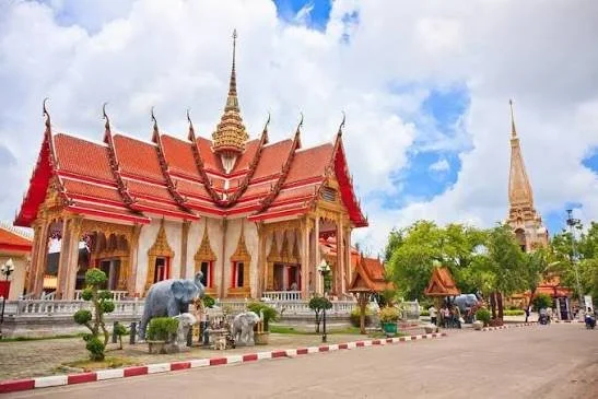 Colorful Thai temple with red and gold roof, surrounded by trees, elephant statues, and a clear sky.