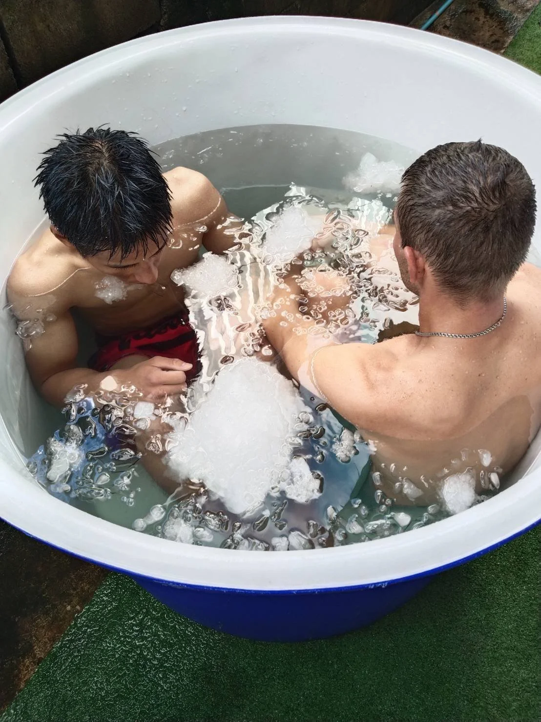 Two men and a boy sitting in a large white plastic tub filled with water and bubbles, outdoors.