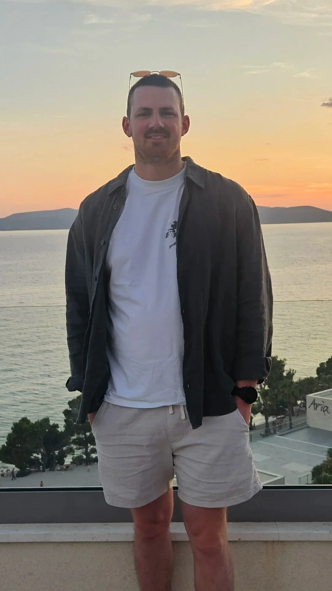 A man standing outdoors near a body of water during sunset, wearing a white t-shirt, beige shorts, a dark jacket, and sunglasses on his head.