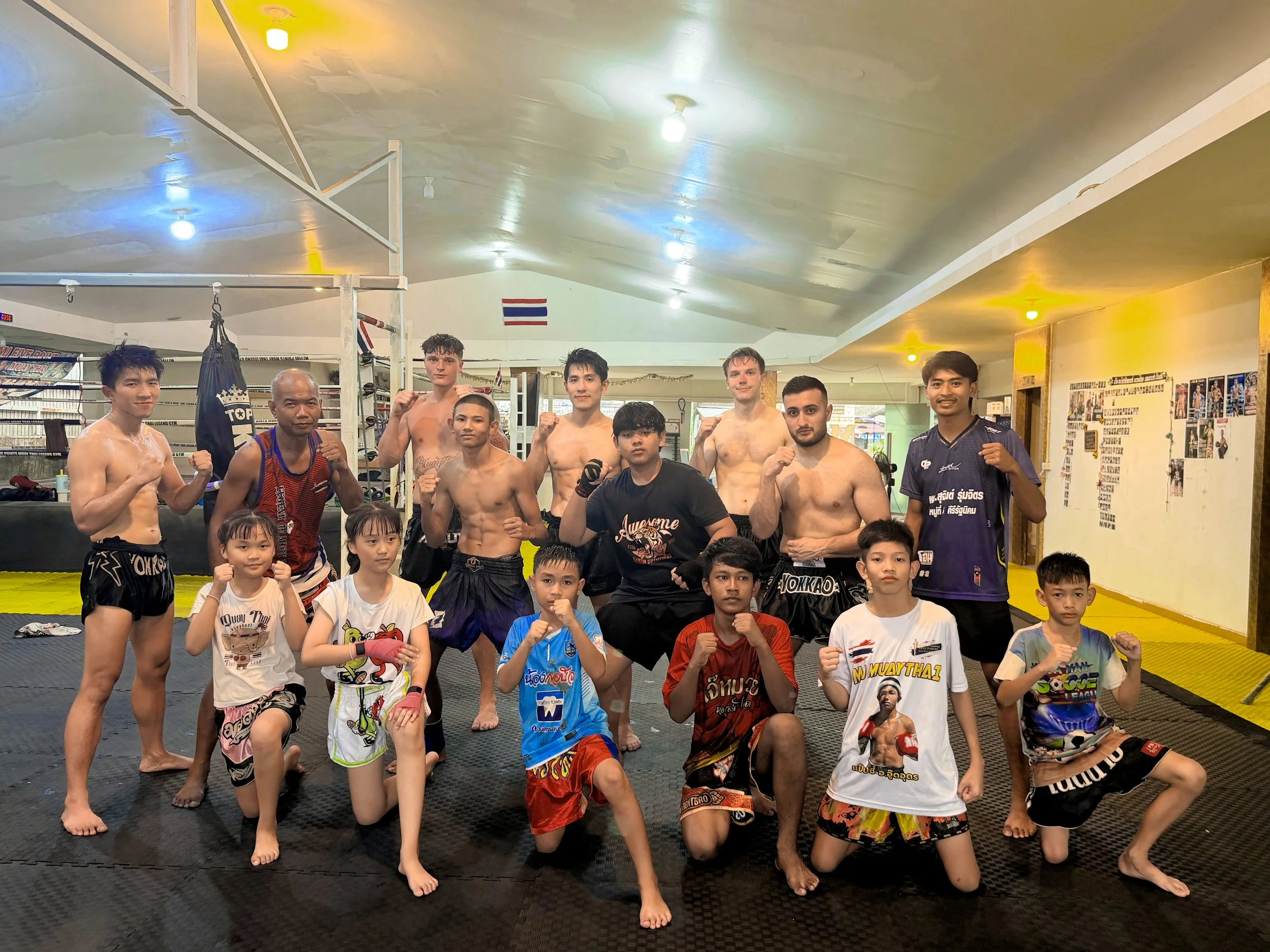 A group of mixed martial artists and children in a martial arts training gym, standing barefoot on a black padded floor, making fists and posing for a photo. The gym has Thai flags and boxing equipment in the background.
