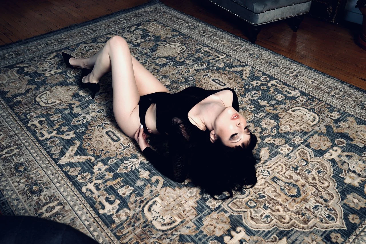 A woman dressed in black lingerie and black high heels lies on her back on a patterned rug in a room with wooden floor and a sofa.