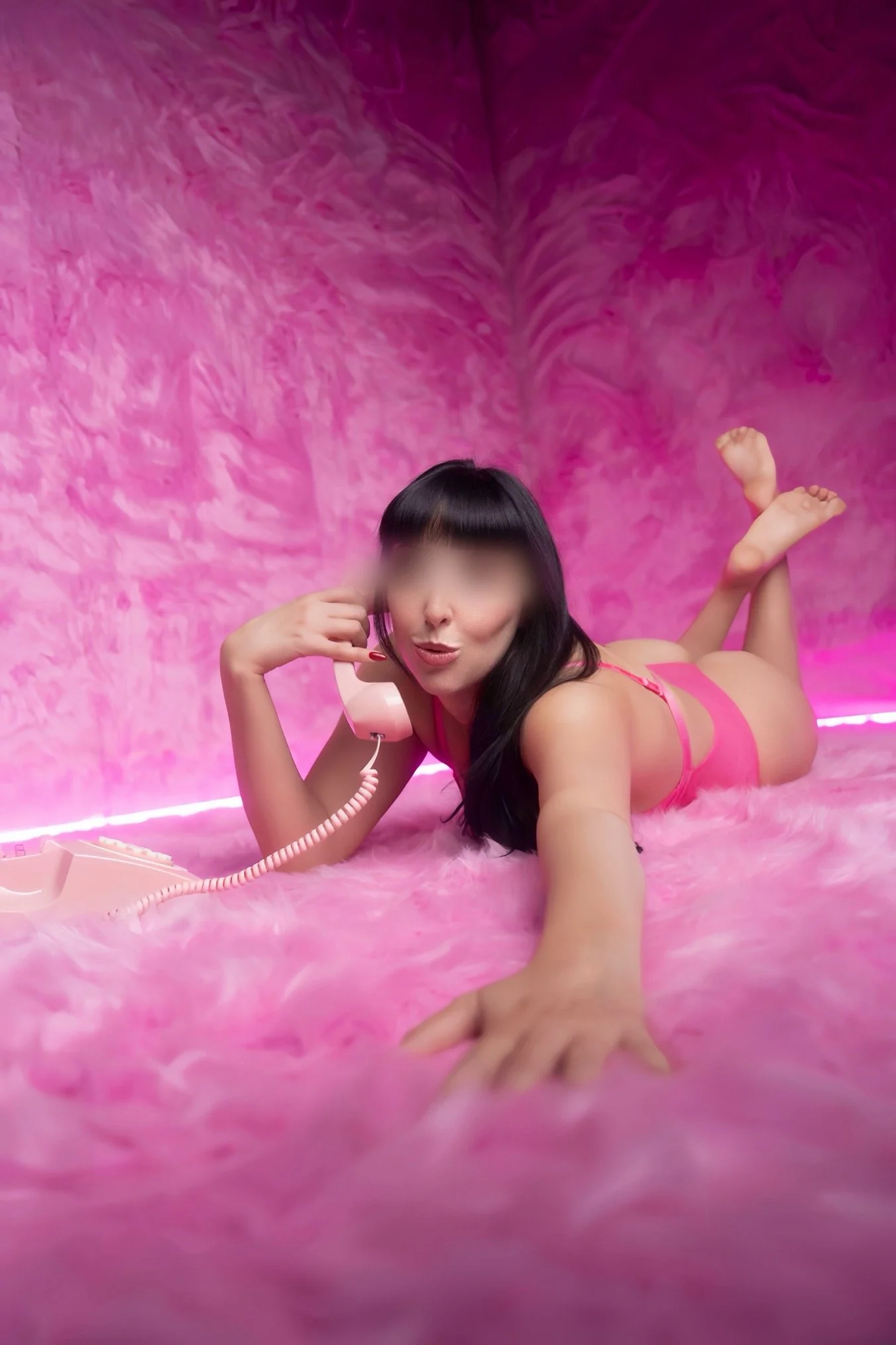 Woman lying on pink fluffy bed, wearing pink lingerie, talking on pink telephone, with a pink wall background.
