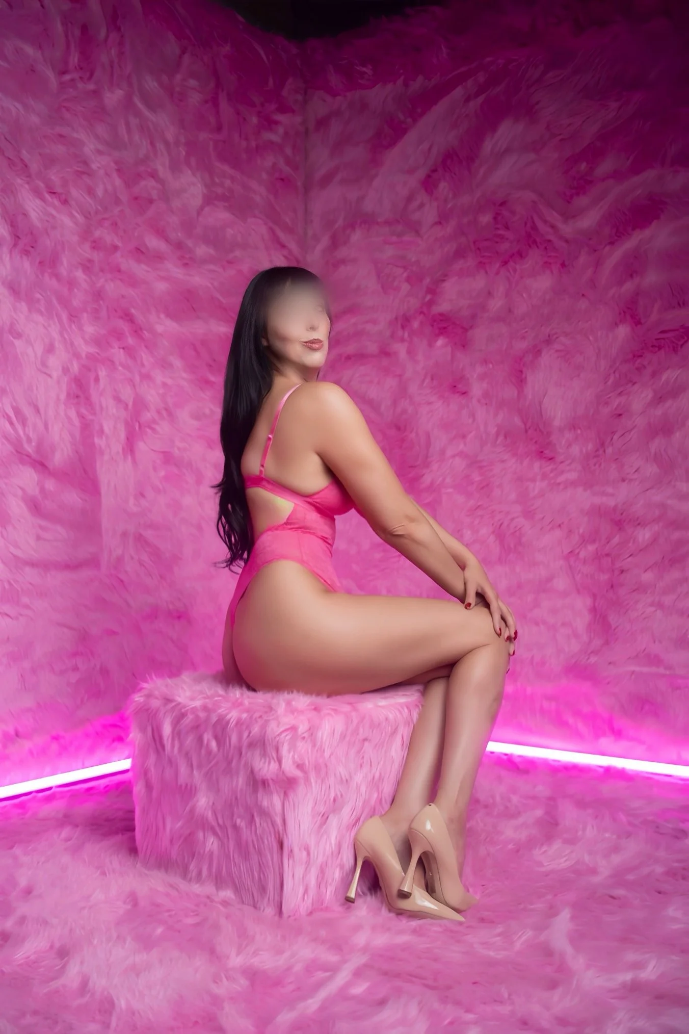 A woman sitting on a pink furry chair in front of a pink furry backdrop, wearing pink lingerie and beige high heels.
