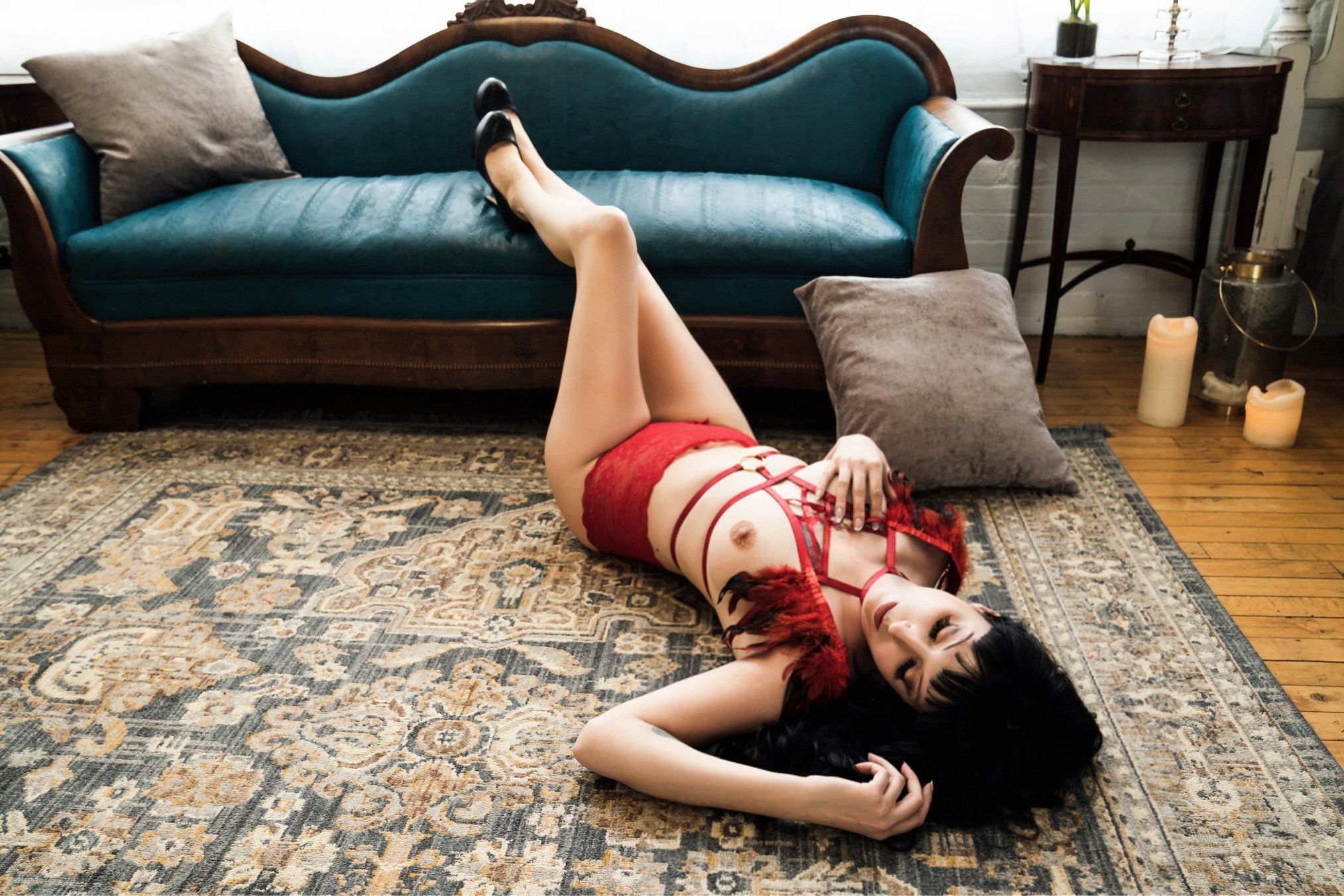 A woman lying on her back on a patterned rug in front of a vintage blue sofa, wearing red lingerie and black shoes, with candles and decorative items on a side table nearby.