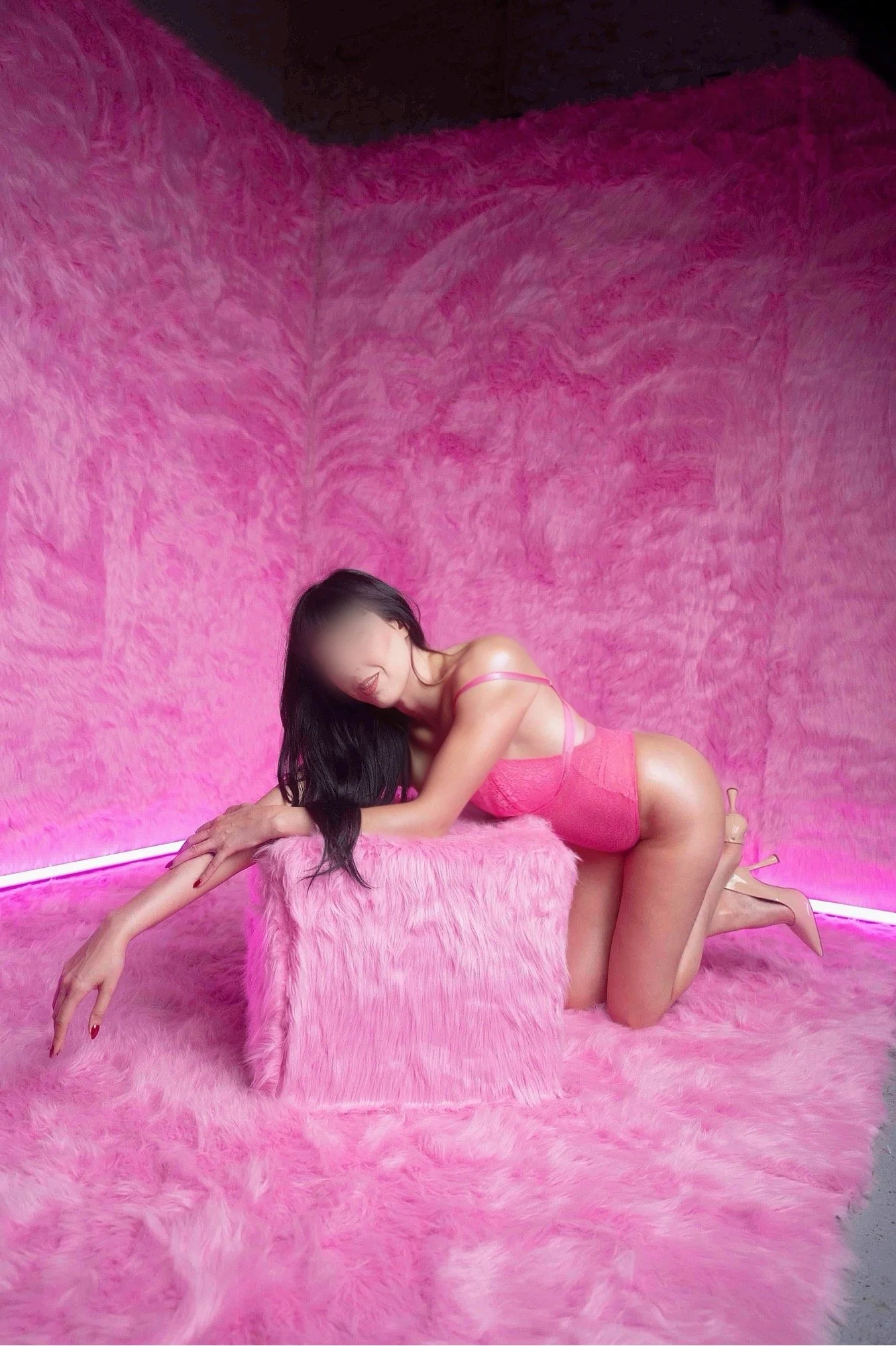 A woman with long black hair and light skin dressed in pink lingerie posing on a pink furry surface with a matching pink furry wall in the background.