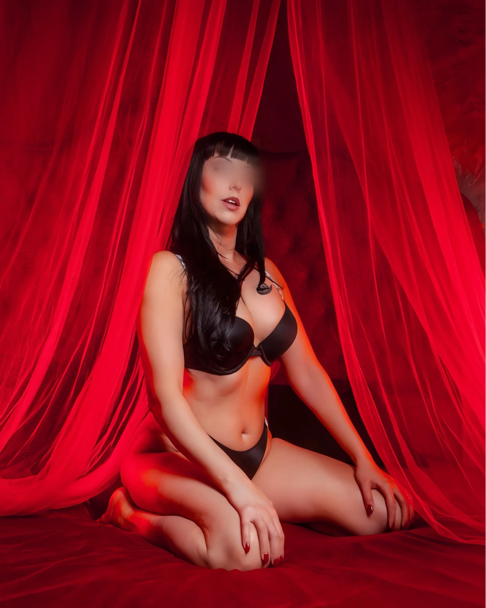 A woman with dark hair and bangs, wearing black lingerie, sitting on a red bedsheet with red curtains behind her, with her eyes blurred for anonymity.