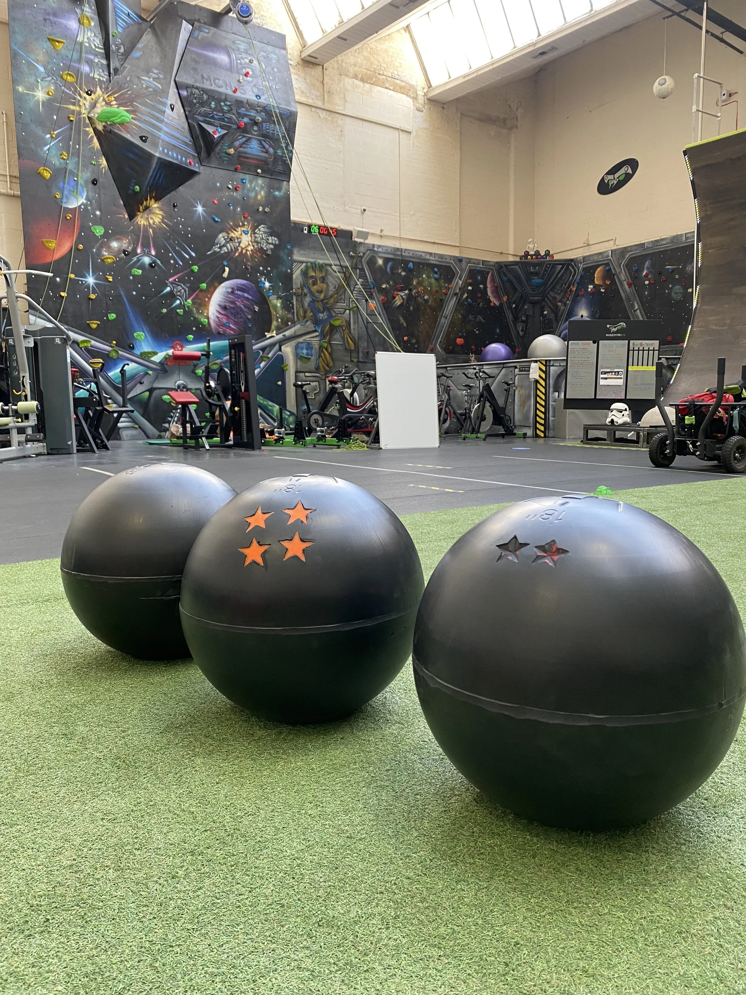 Three black exercise balls with star cutouts, placed on a green artificial turf at an indoor climbing gym with colorful mural walls depicting space and cosmic themes, and various climbing and workout equipment in the background.