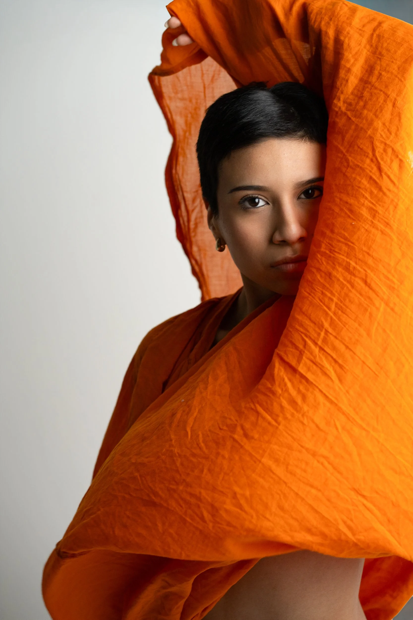 Amanda, portrait session at 9 Studios, SoHo NYC — face emerging through draped orange linen fabric held overhead, steady gaze, white background, May 2026