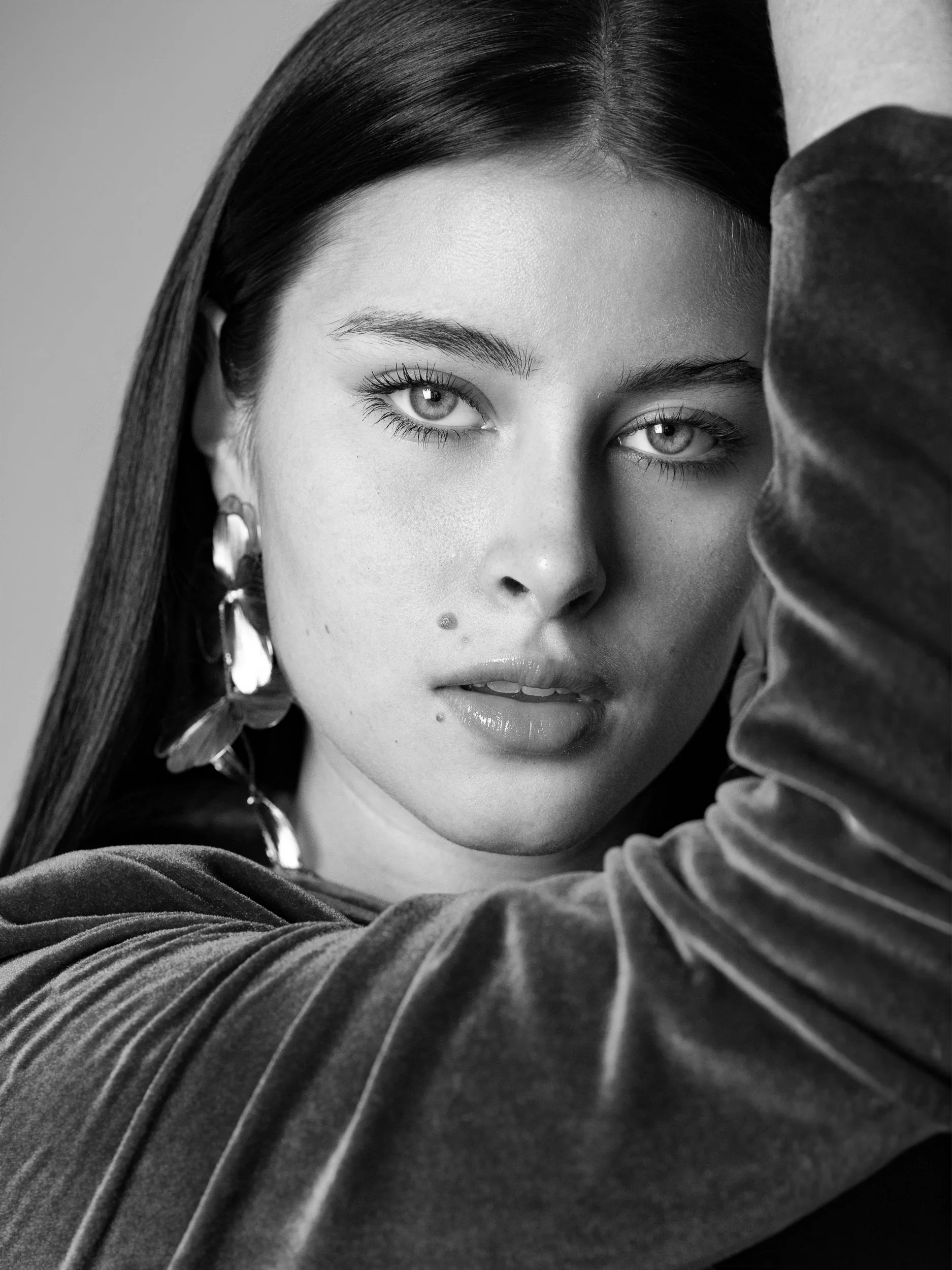 Gloria, close-up black and white portrait with direct gaze, statement earrings, velvet top, arm raised, studio