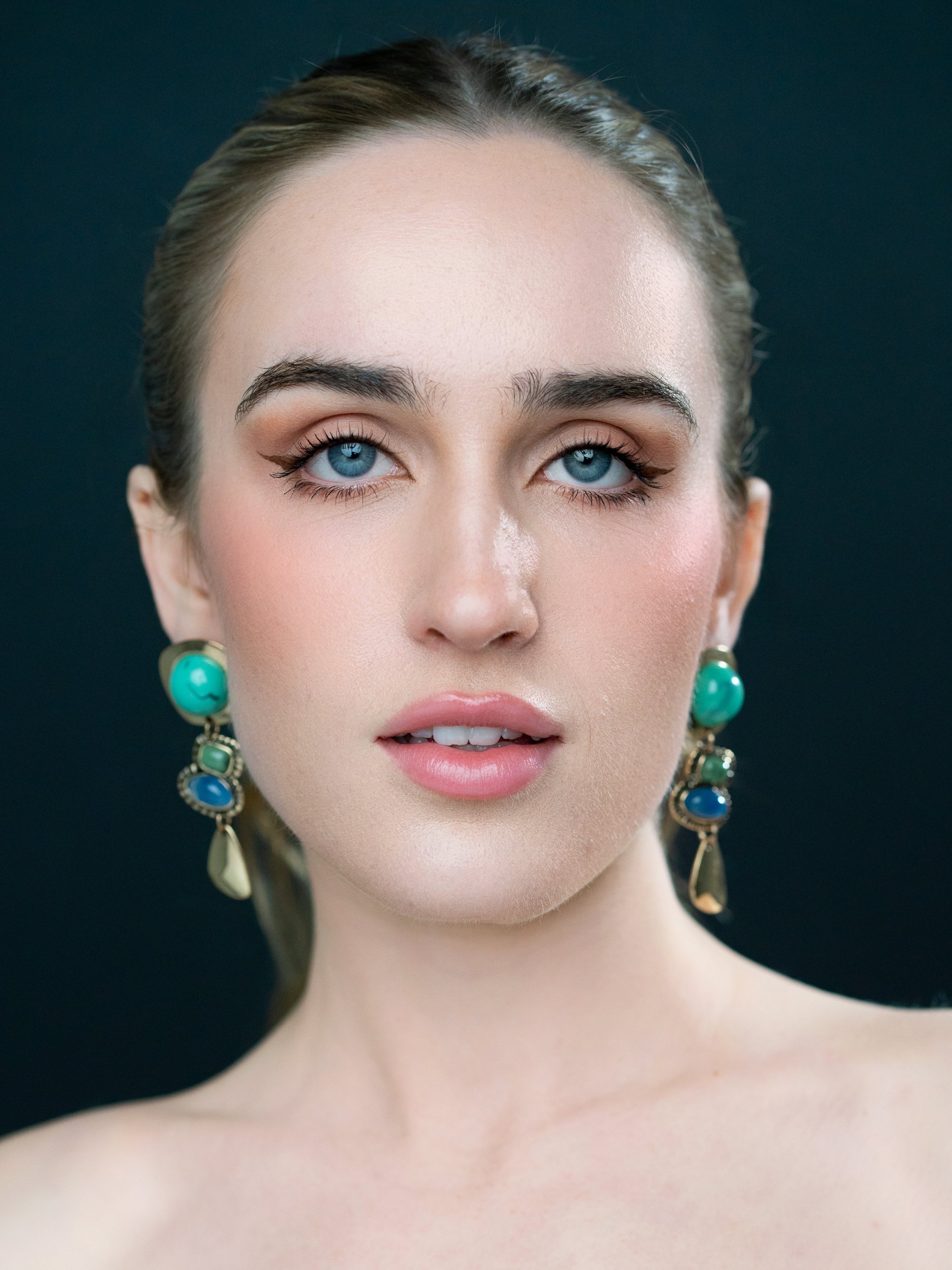 Kyra in a symmetric beauty portrait, luminous skin and pale blue eyes, ornate turquoise-and-gold drop earrings, deep teal studio backdrop.