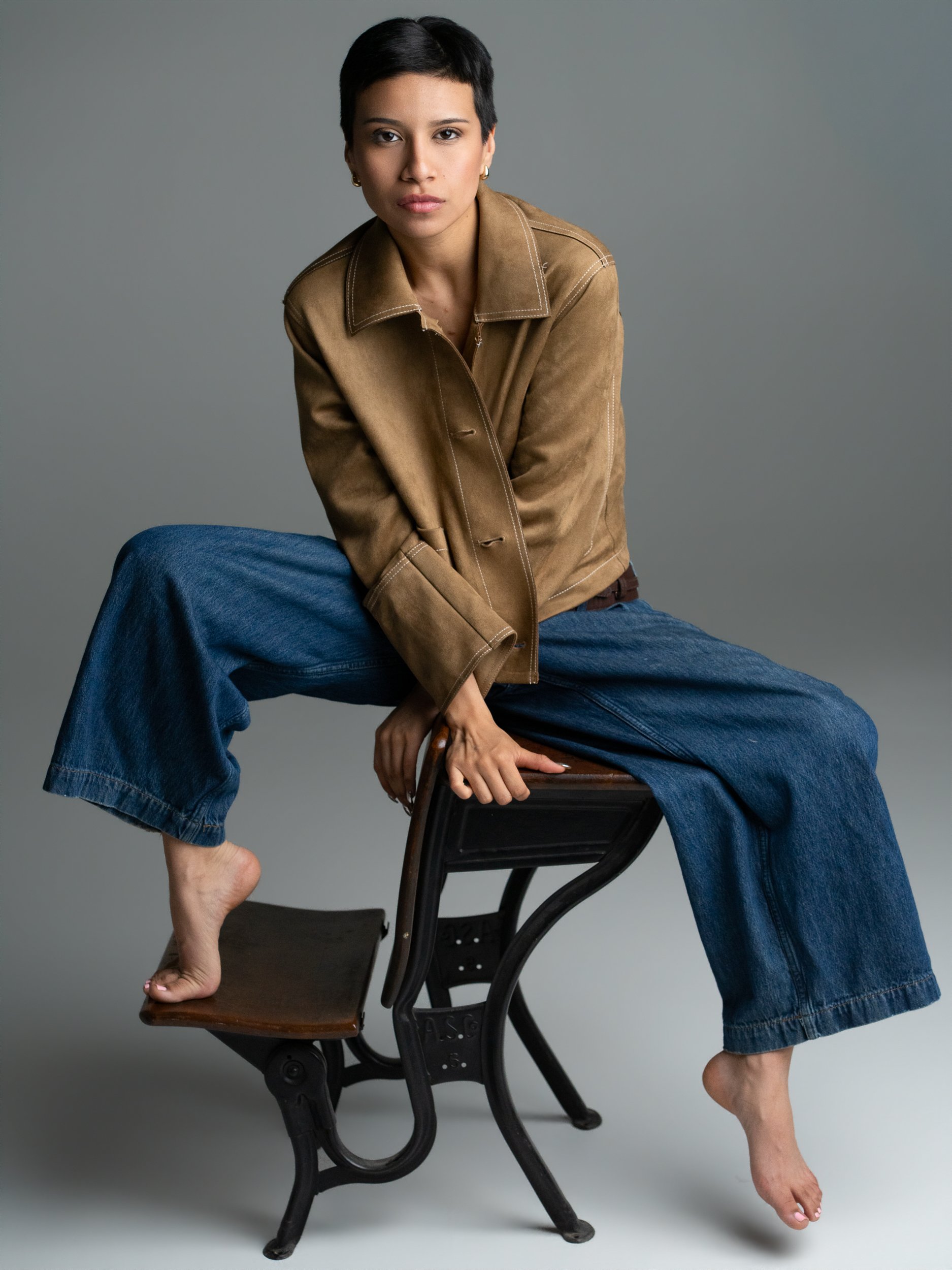 Amanda, portrait session at 9 Studios, SoHo NYC — full-length editorial, seated on vintage iron chair, tan suede jacket, wide-leg denim, barefoot, direct gaze, gray background, May 2026