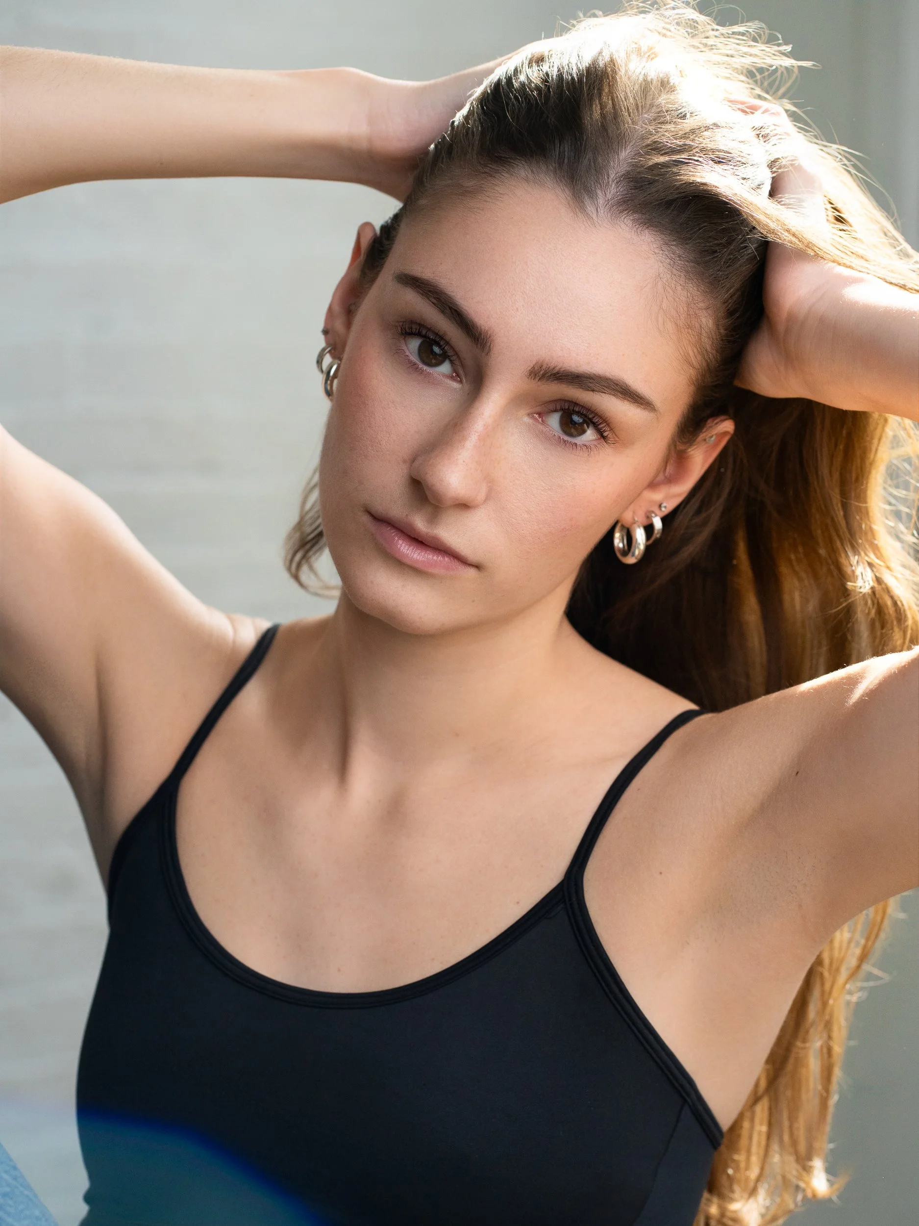 Young woman with long light brown hair holding her hands to her head, wearing a black tank top and silver hoop earrings, looking at the camera in warm natural light.