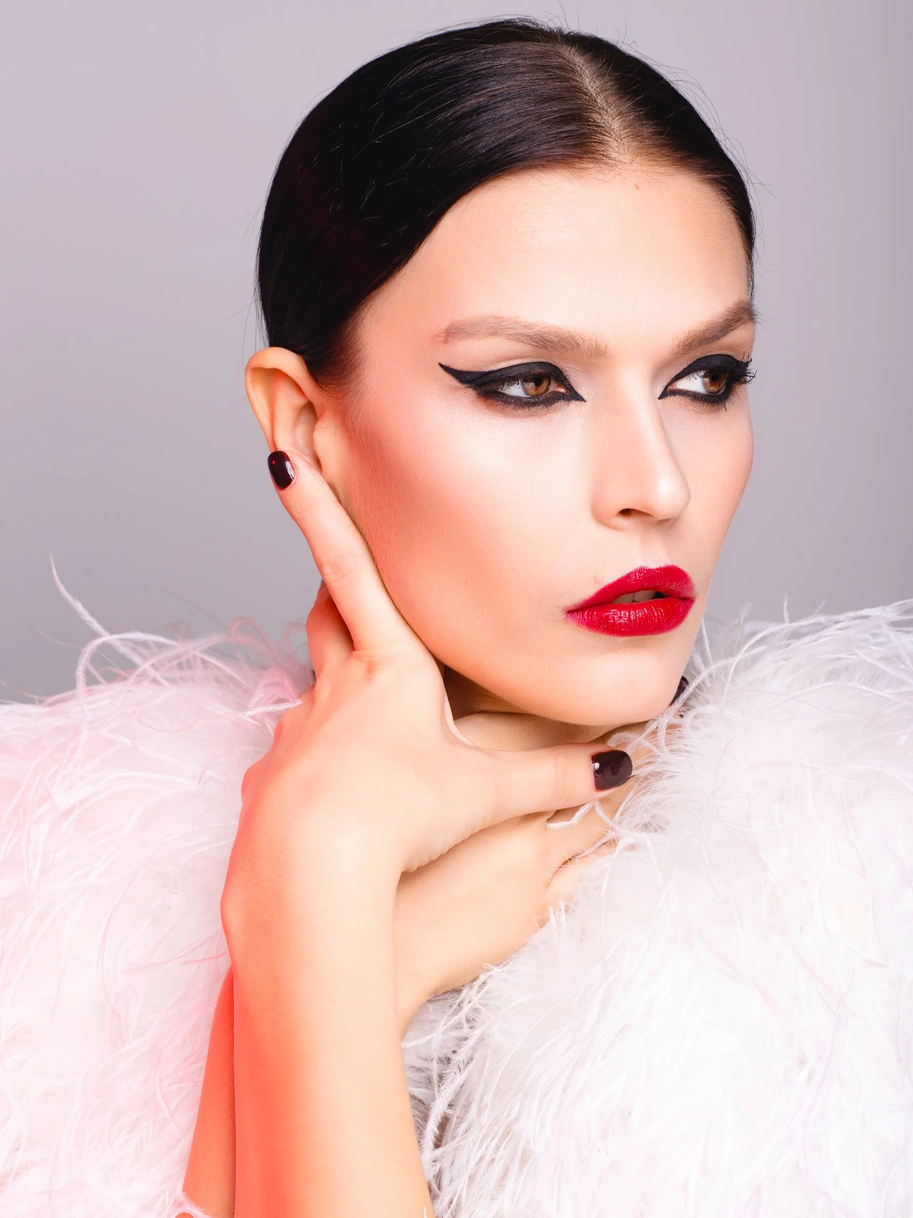 Lisa gazing off-camera with a cool expression, slicked-back hair, bold graphic eyeliner and glossy red lips, plush white feather boa, soft gray studio.