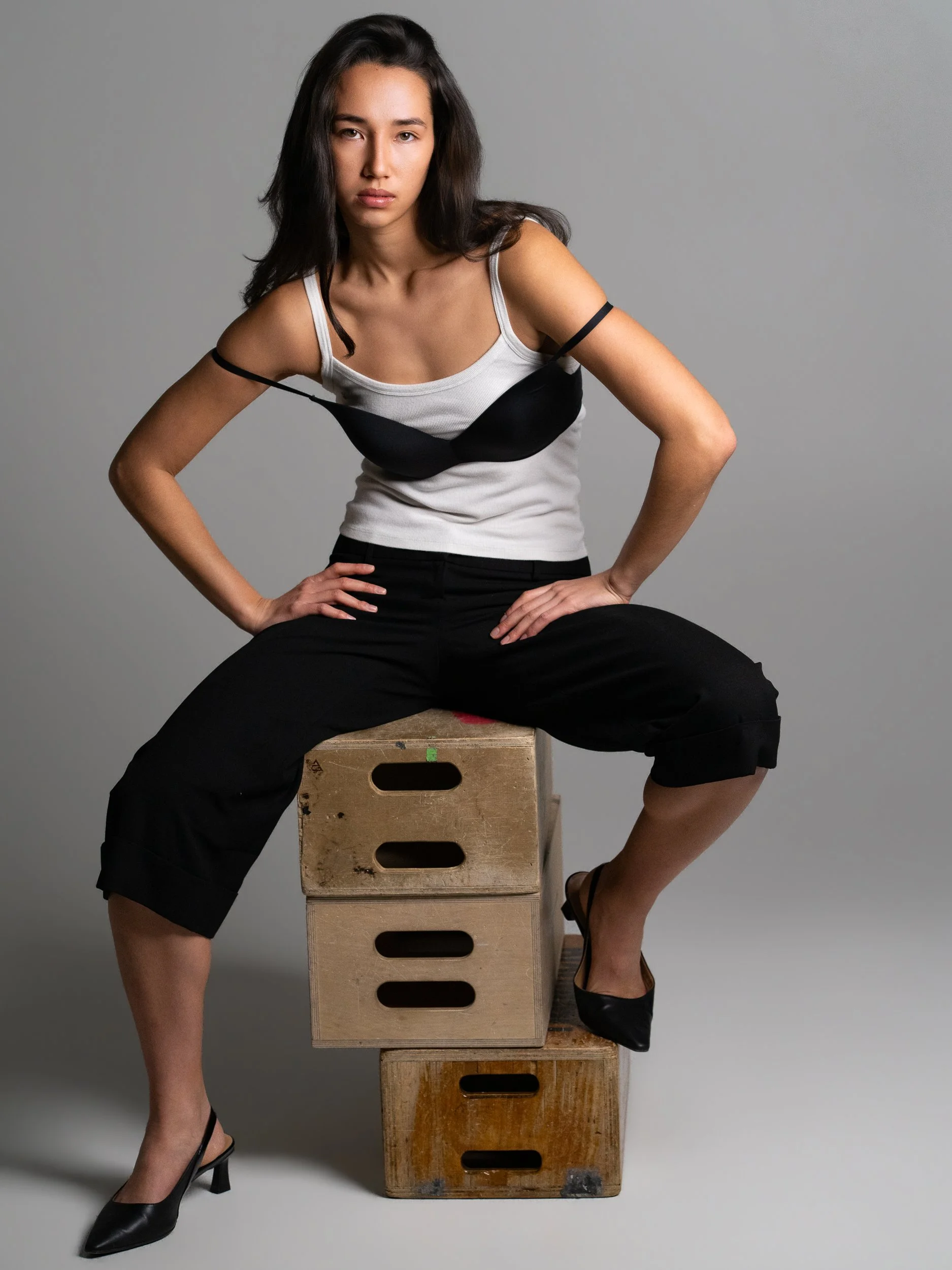 Sophie, seated on apple crates wearing black bra over white tank top, direct gaze, studio
