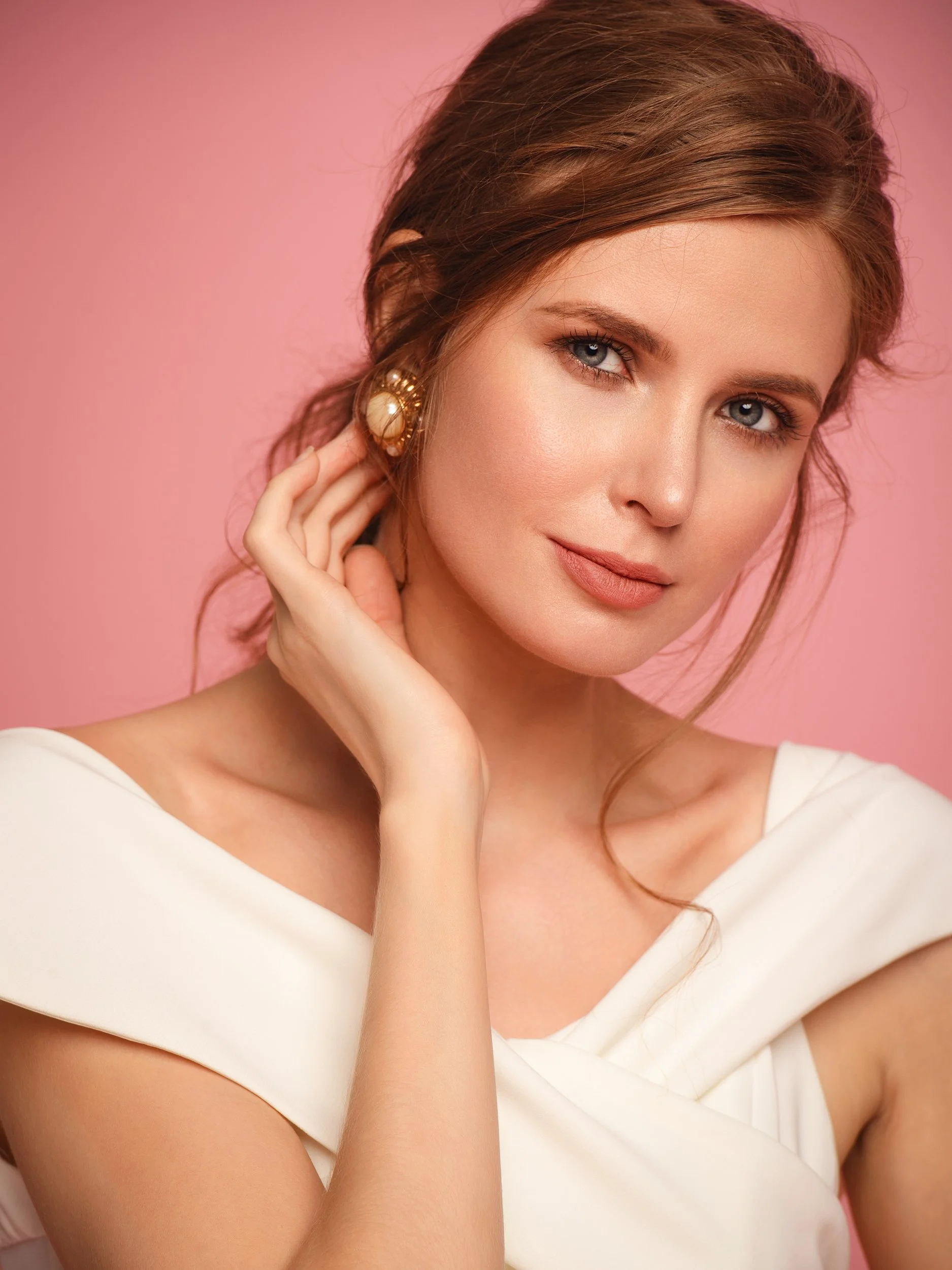Irina with a poised, level gaze and hand resting at her neck, soft updo and gold-and-pearl earring, cream off-shoulder dress, blush-pink studio.