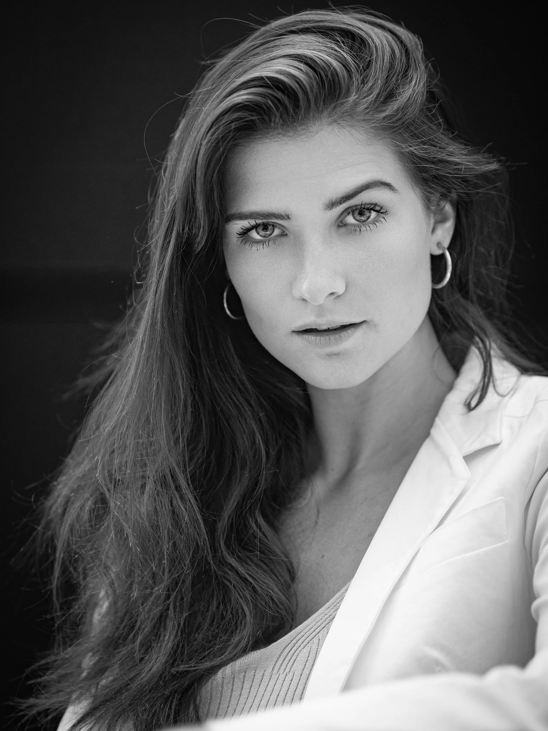 Emma, black and white portrait with direct gaze, long wavy hair, white blazer, hoop earrings, dark studio background