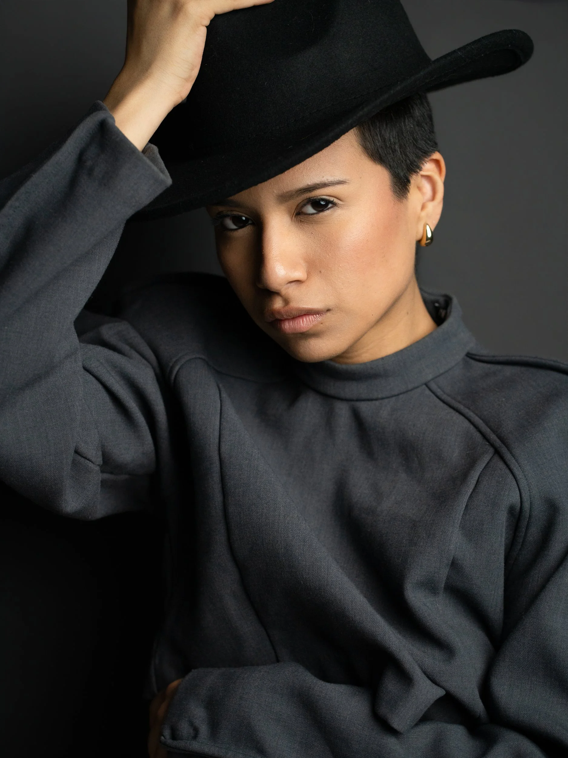 Amanda, portrait session at 9 Studios, SoHo NYC — close editorial portrait, hand pressing wide-brim black hat down, sculpted-shoulder gray jacket, steady under-brim gaze, dark gray background, May 2026