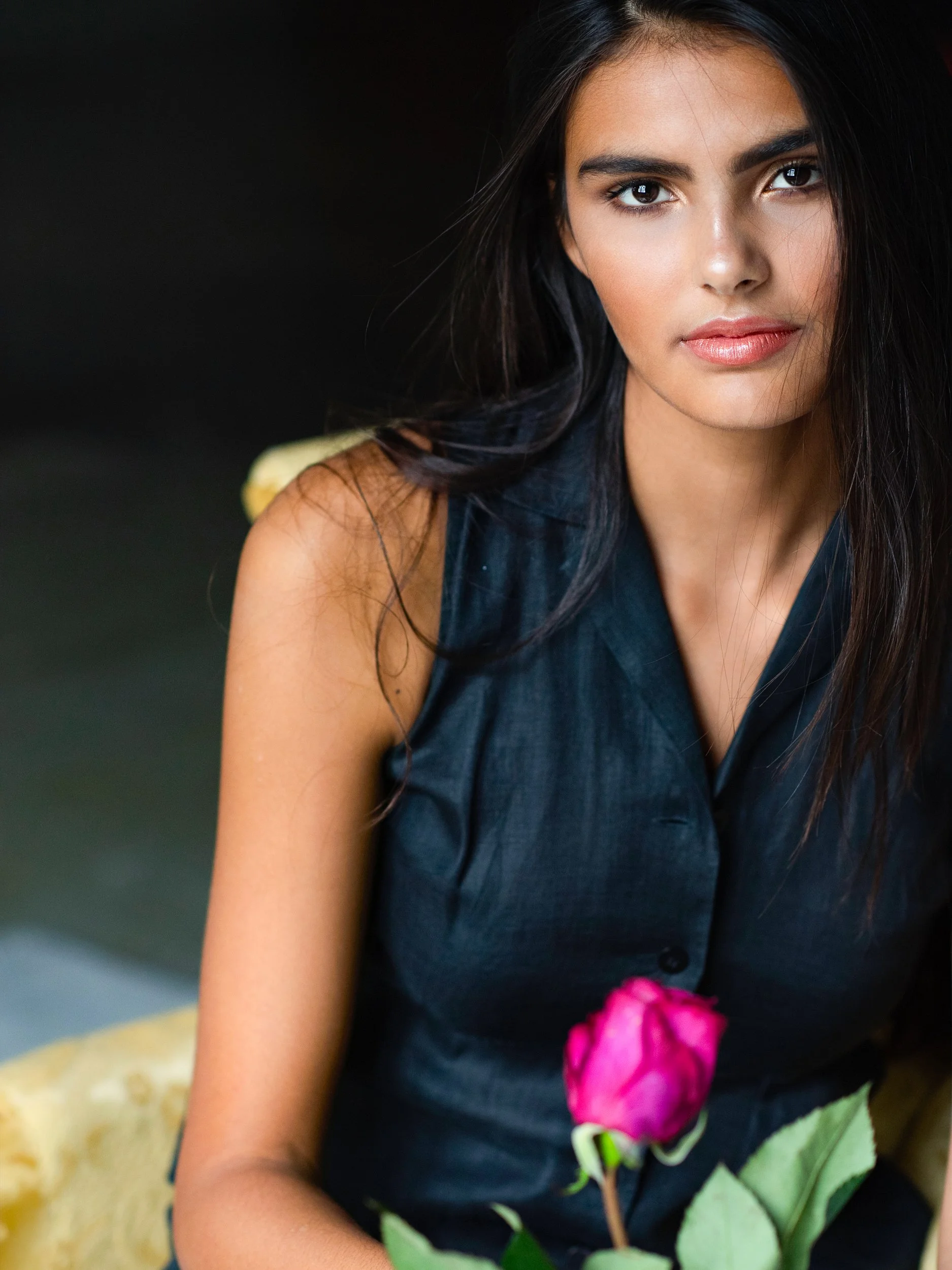 Mya looking past the camera in warm window light, long dark hair and black blouse, a single magenta rose held in the foreground, deep shadowed setting.