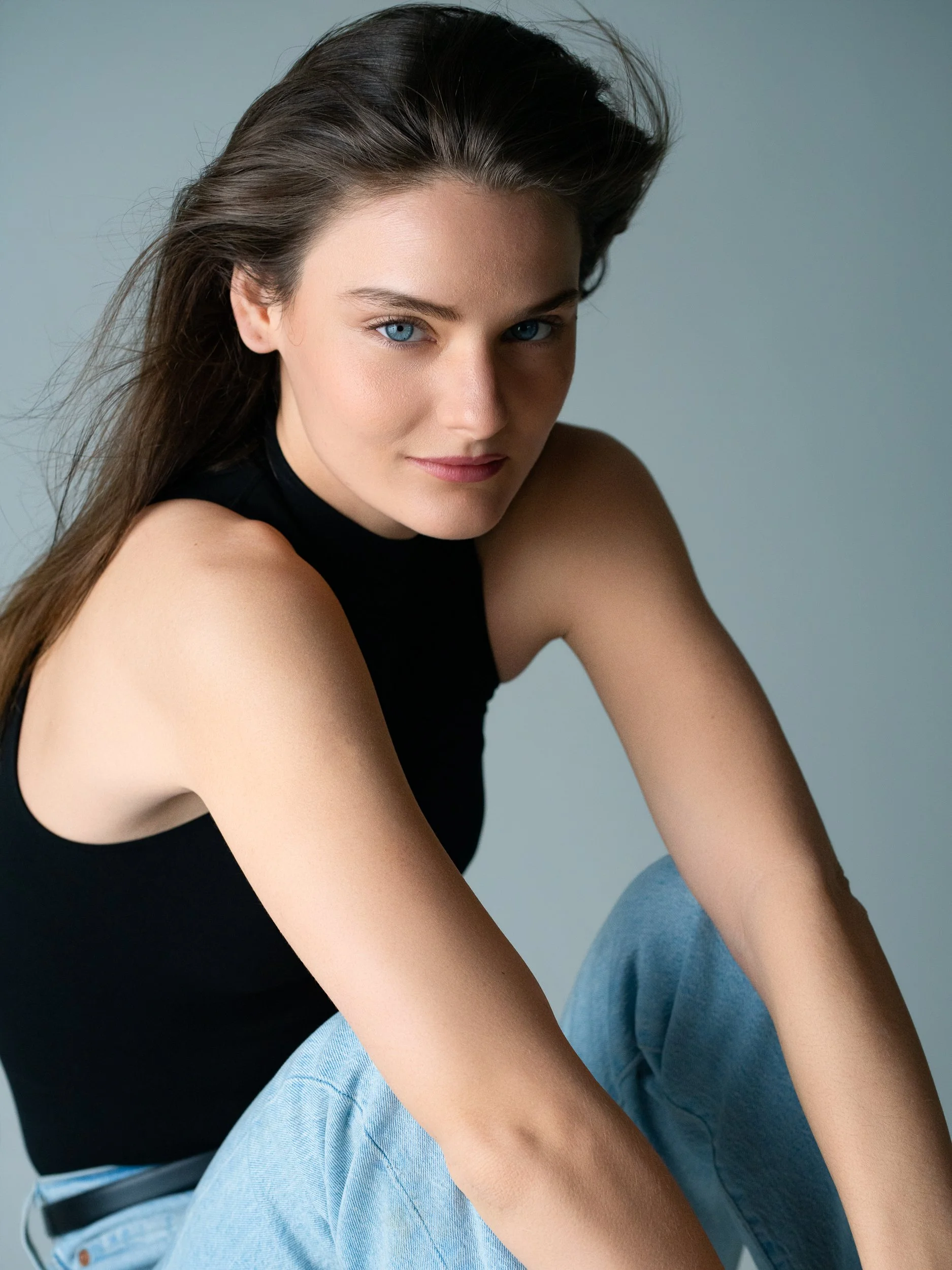 Lucy with chin slightly lowered, long brown hair and pale blue eyes, black mock-neck top and denim, muted studio backdrop.