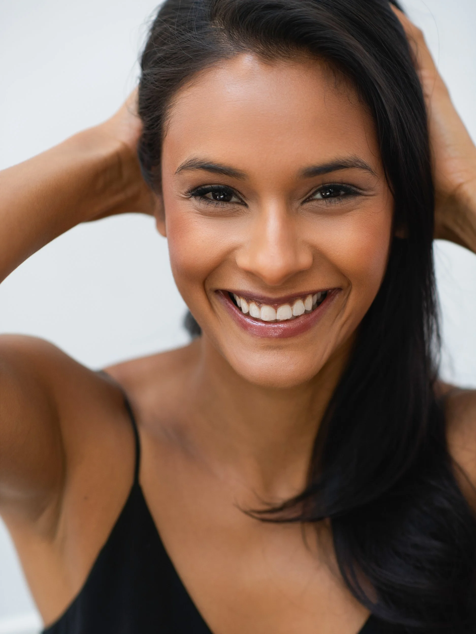 Didi laughing toward the camera, hands swept up into her dark hair, natural makeup and black slip top, light, airy studio.
