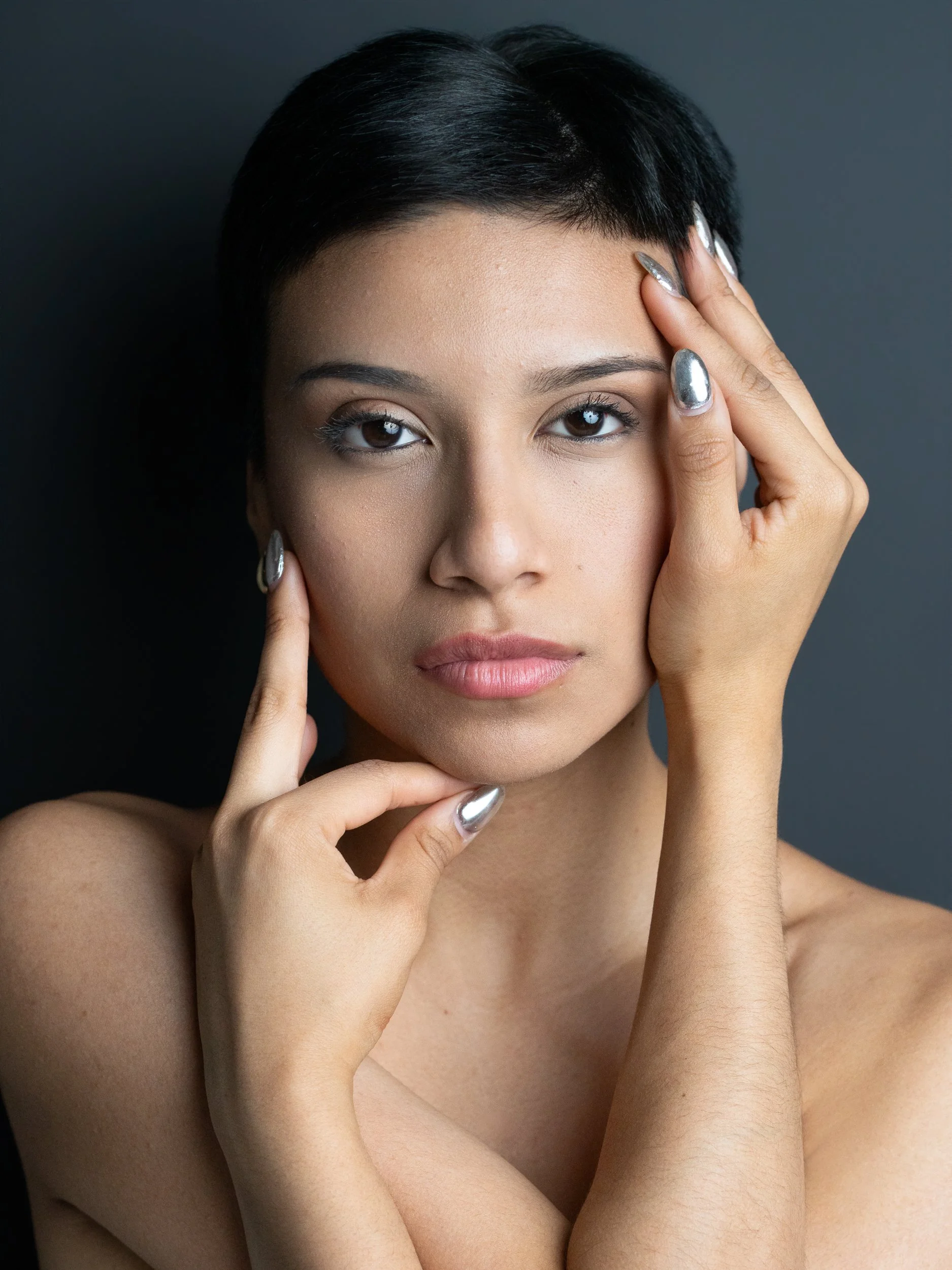 Amanda, portrait session at 9 Studios, SoHo NYC — close beauty portrait, both hands framing face, chrome silver nails, bare shoulders, steady gaze, dark gray background, May 2026