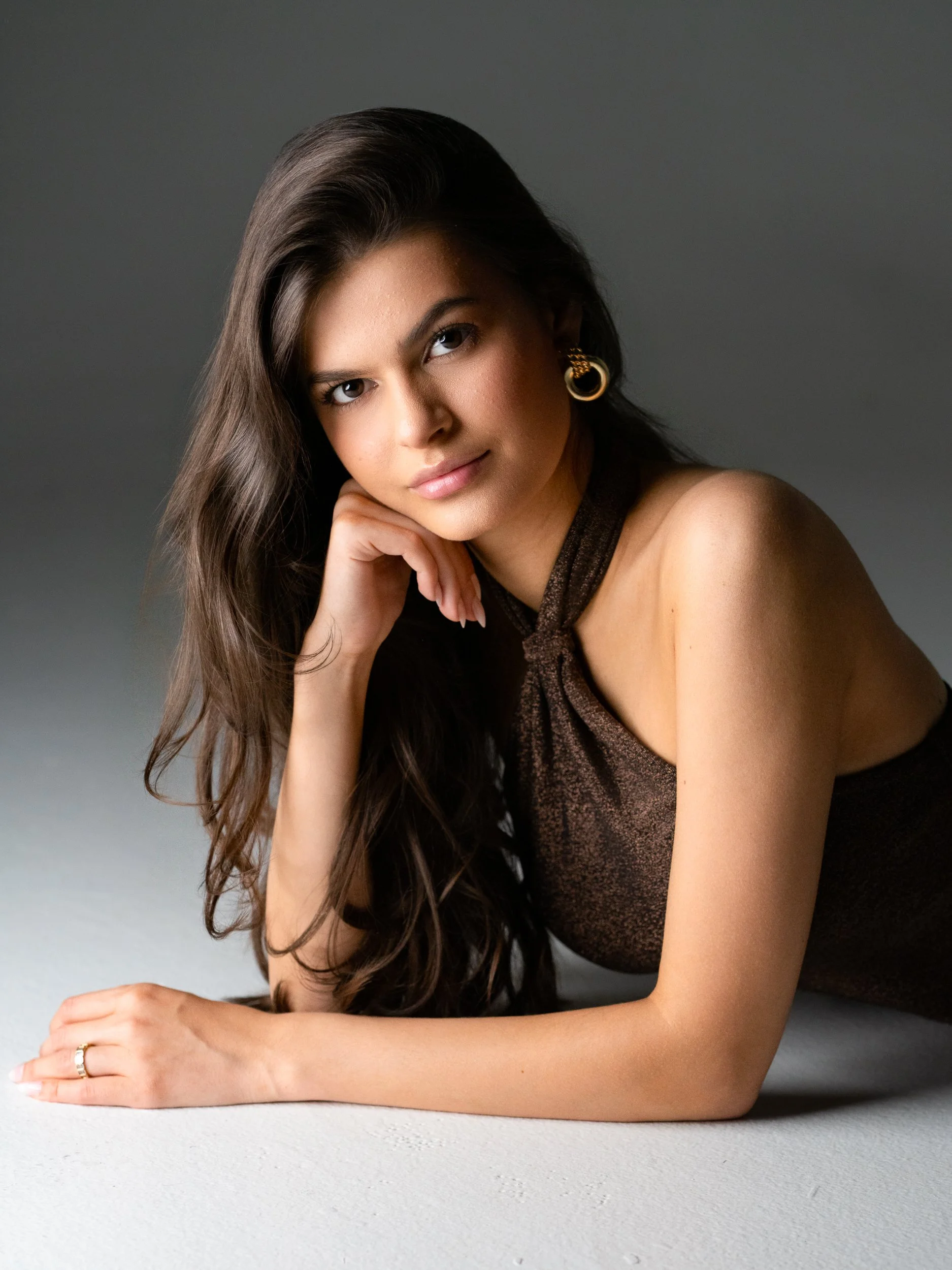 Elisa reclining on the floor, gaze direct to camera, long wavy hair, shimmering brown halter and gold hoop earrings, moody studio light.