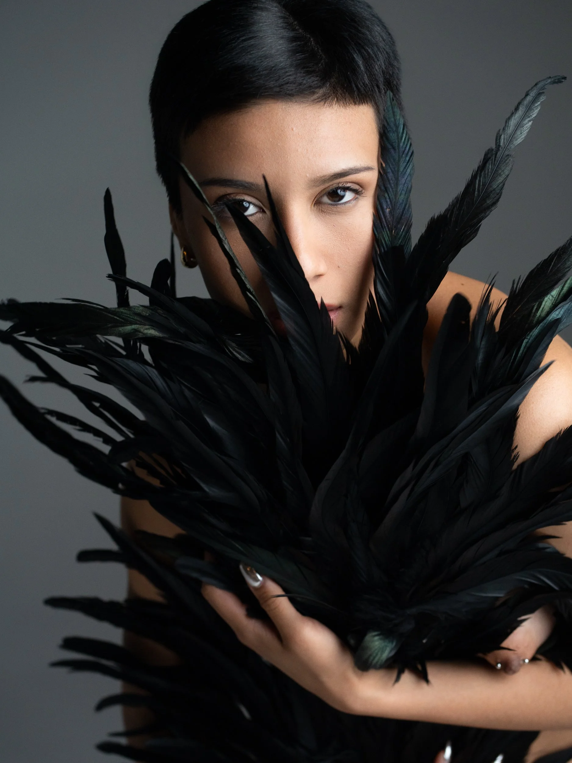 Amanda, portrait session at 9 Studios, SoHo NYC — half-face obscured by iridescent black feathers held close, one eye and bare shoulder visible against neutral gray, May 2026