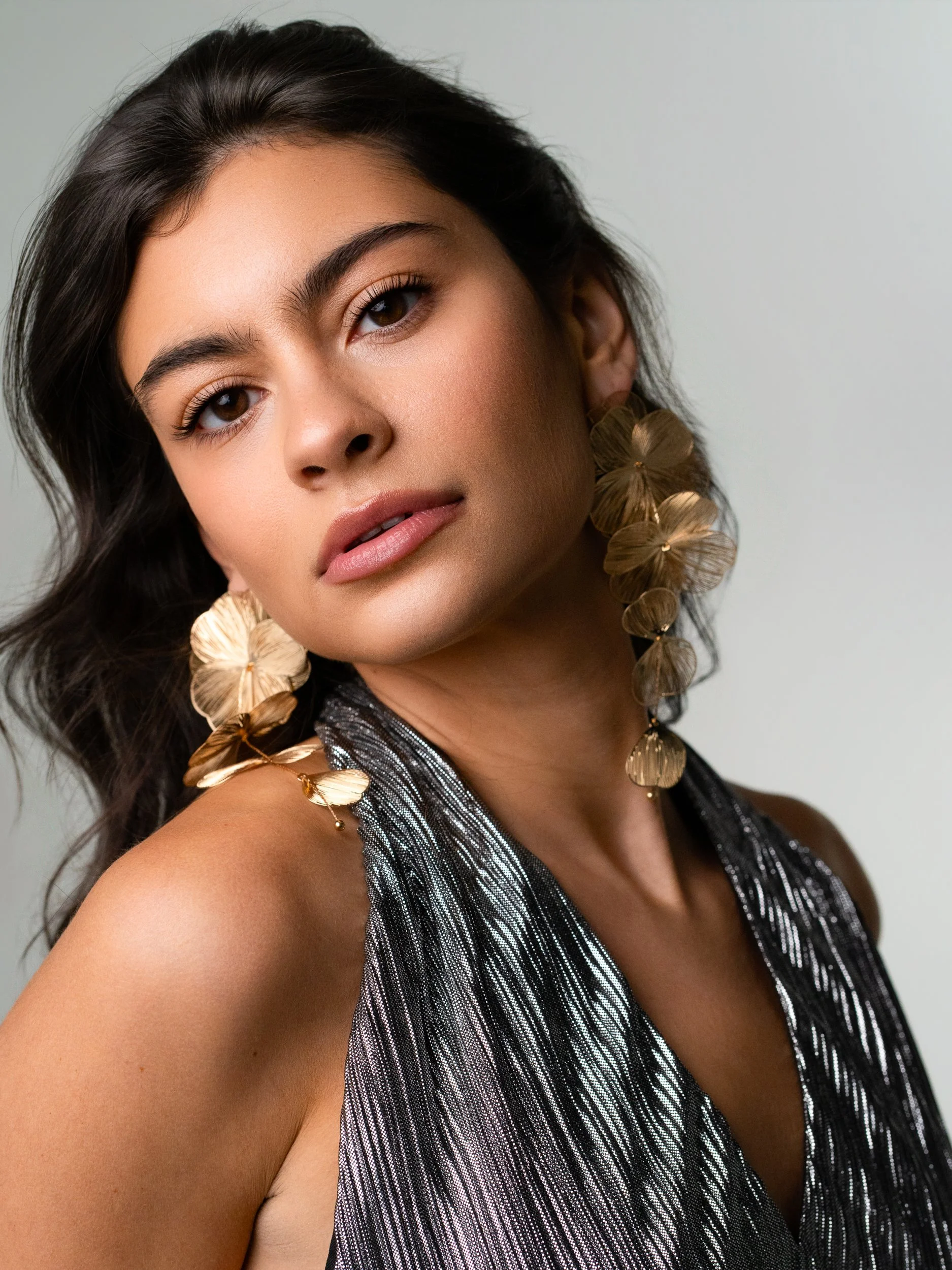 Hannah with a cool, level gaze turned over her shoulder, dark waves and sculptural gold flower earrings, shimmering silver halter, soft gray studio.
