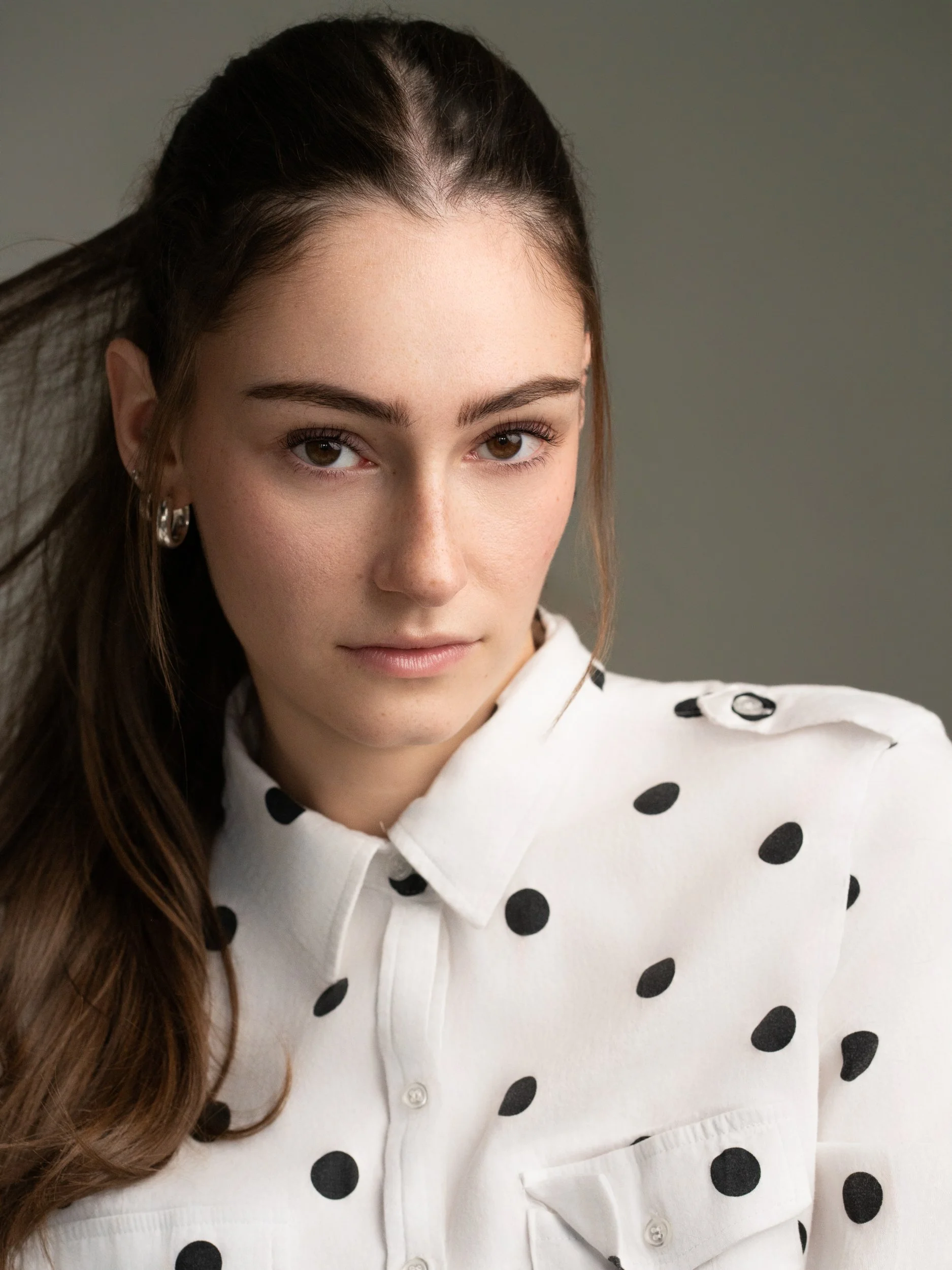 Young woman with long dark hair looking at the camera with a calm expression, wearing a white polka-dot shirt and silver hoop earrings, against a soft gray-green background.