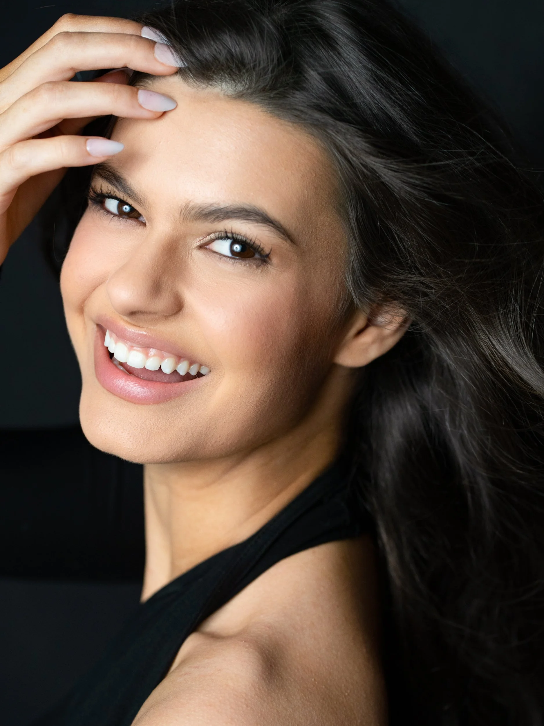 Close beauty portrait of a smiling brunette model against a black backdrop, hand in her hair, glancing to camera.