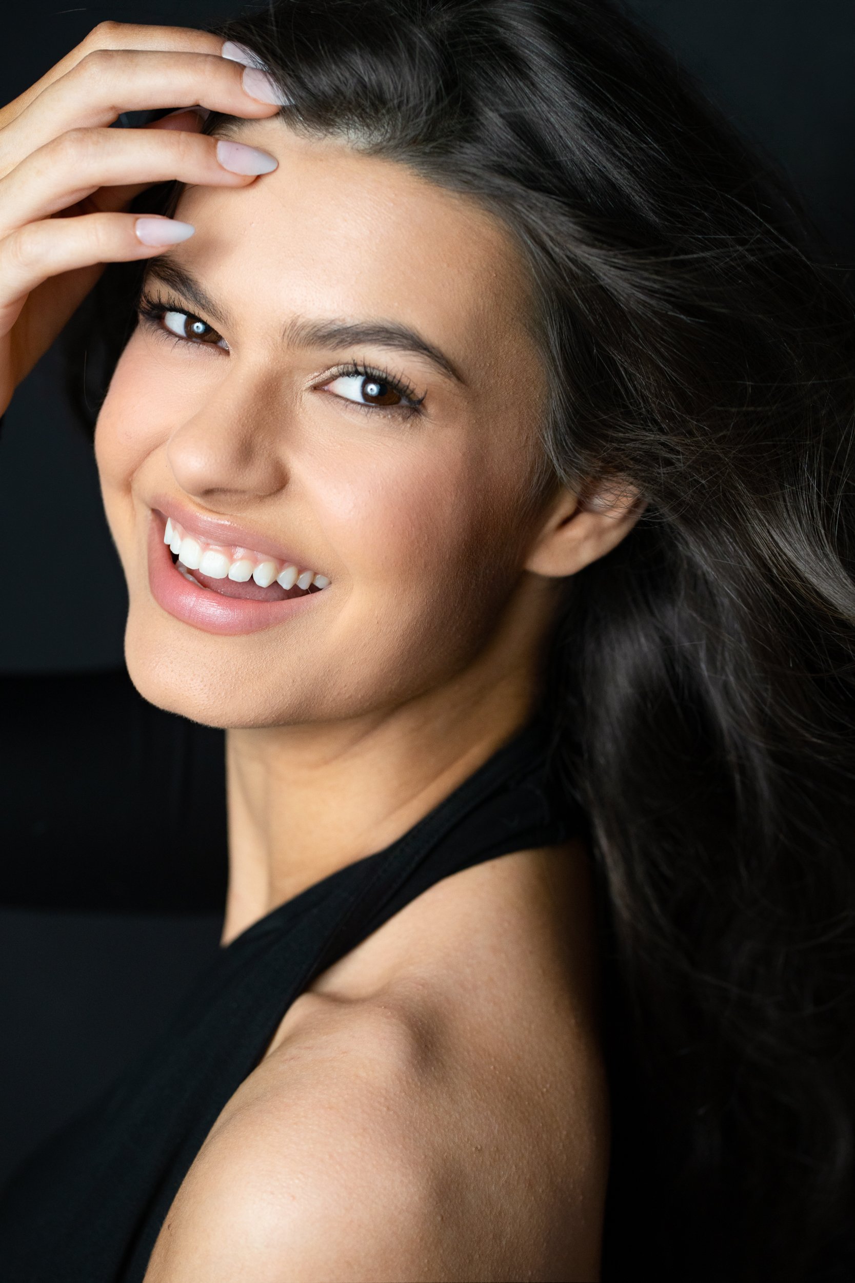 Elisa glancing back with a bright smile, fingers sweeping through her dark hair, black top, low-key studio against black.