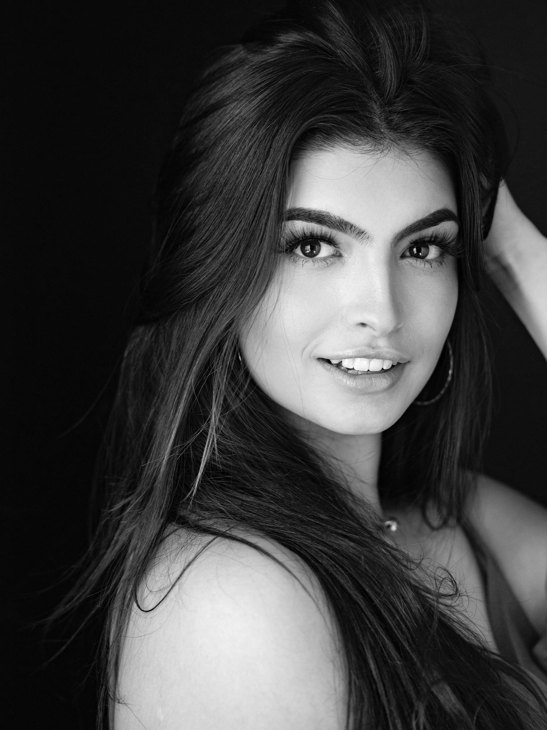Lyssi, black and white portrait with confident smile, long dark hair, hand raised, hoop earring, dark studio background