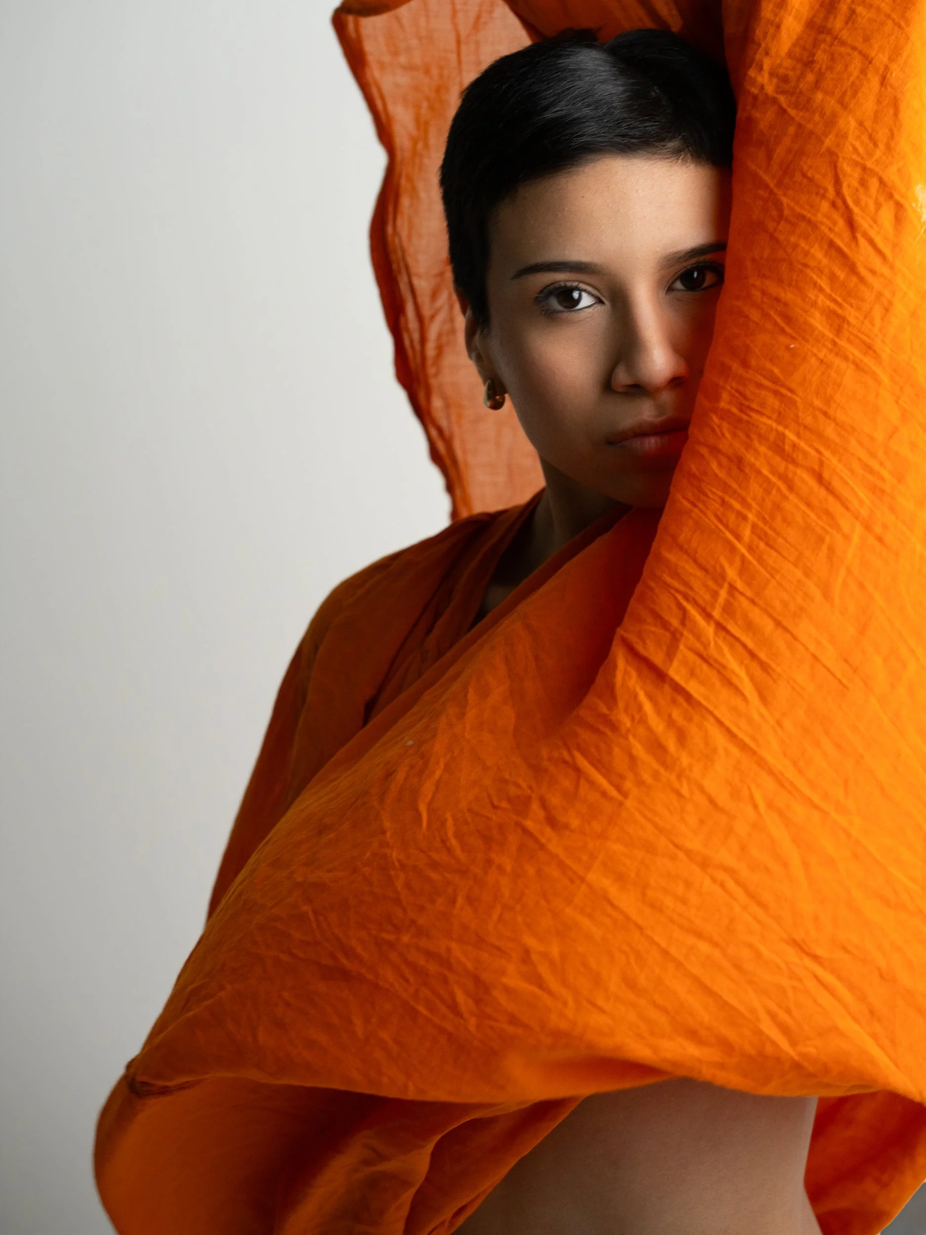 Amanda with a short dark pixie cut, direct gaze to camera, framed by flowing bright orange fabric, soft gray studio.