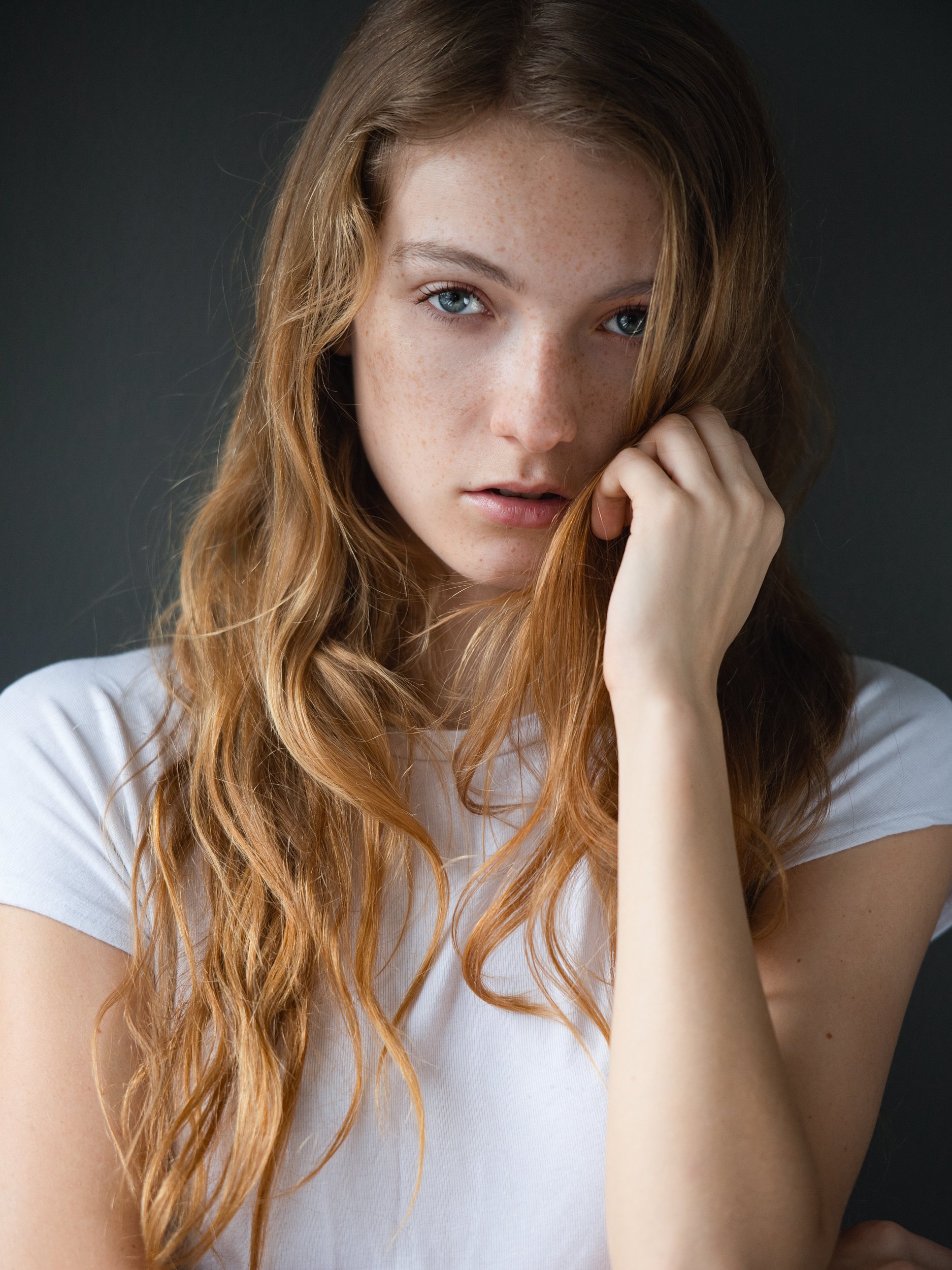 Lauren with a still, pensive gaze, freckled skin and natural strawberry-blonde waves, hand resting in her hair, plain white tee, soft natural light.