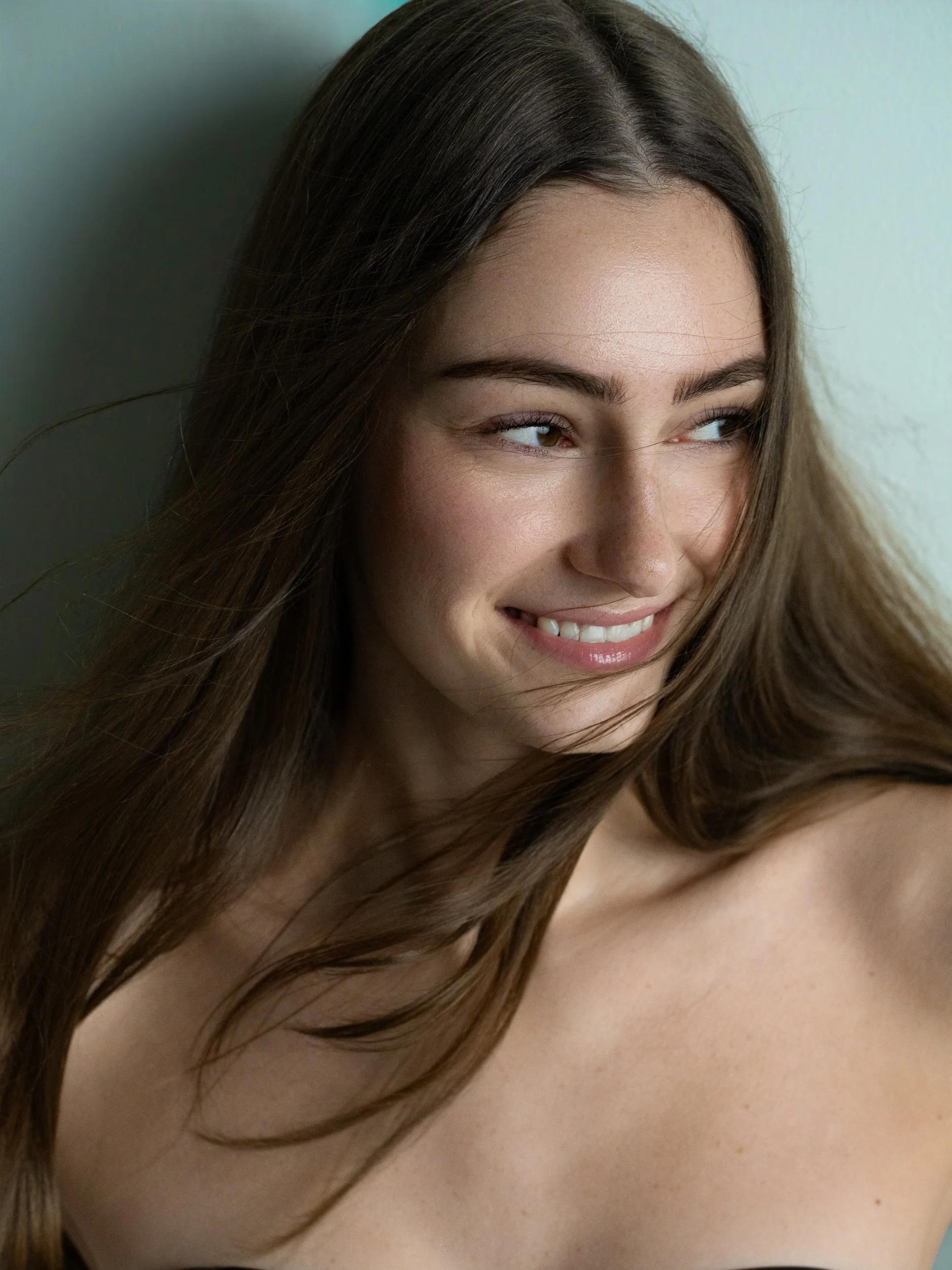 Young woman with long brown hair smiling and looking to the side, bare shoulders, against a soft blue-green background.