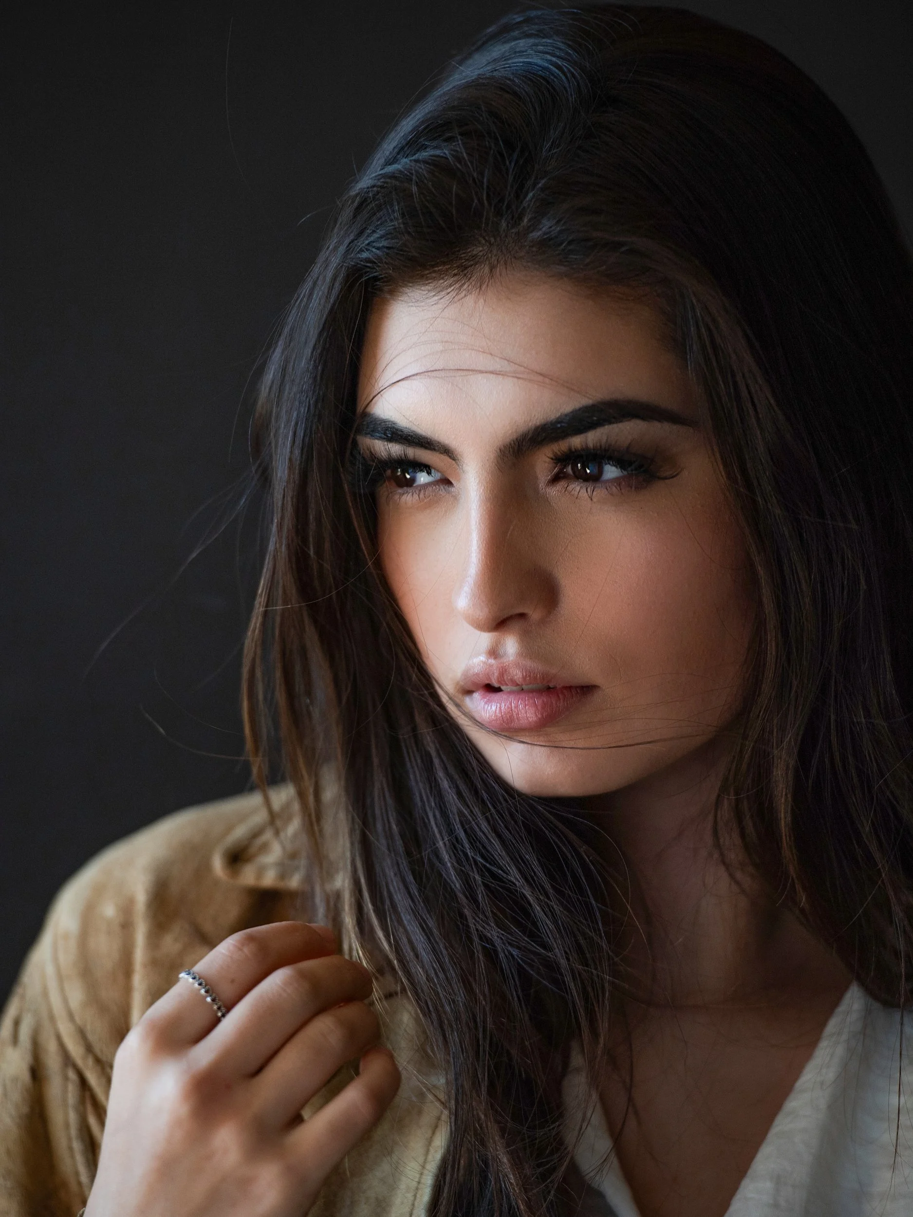 Lyssi with a still, distant gaze, dark hair drifting across her face, hand to a strand, tan suede coat, low-key moody studio.