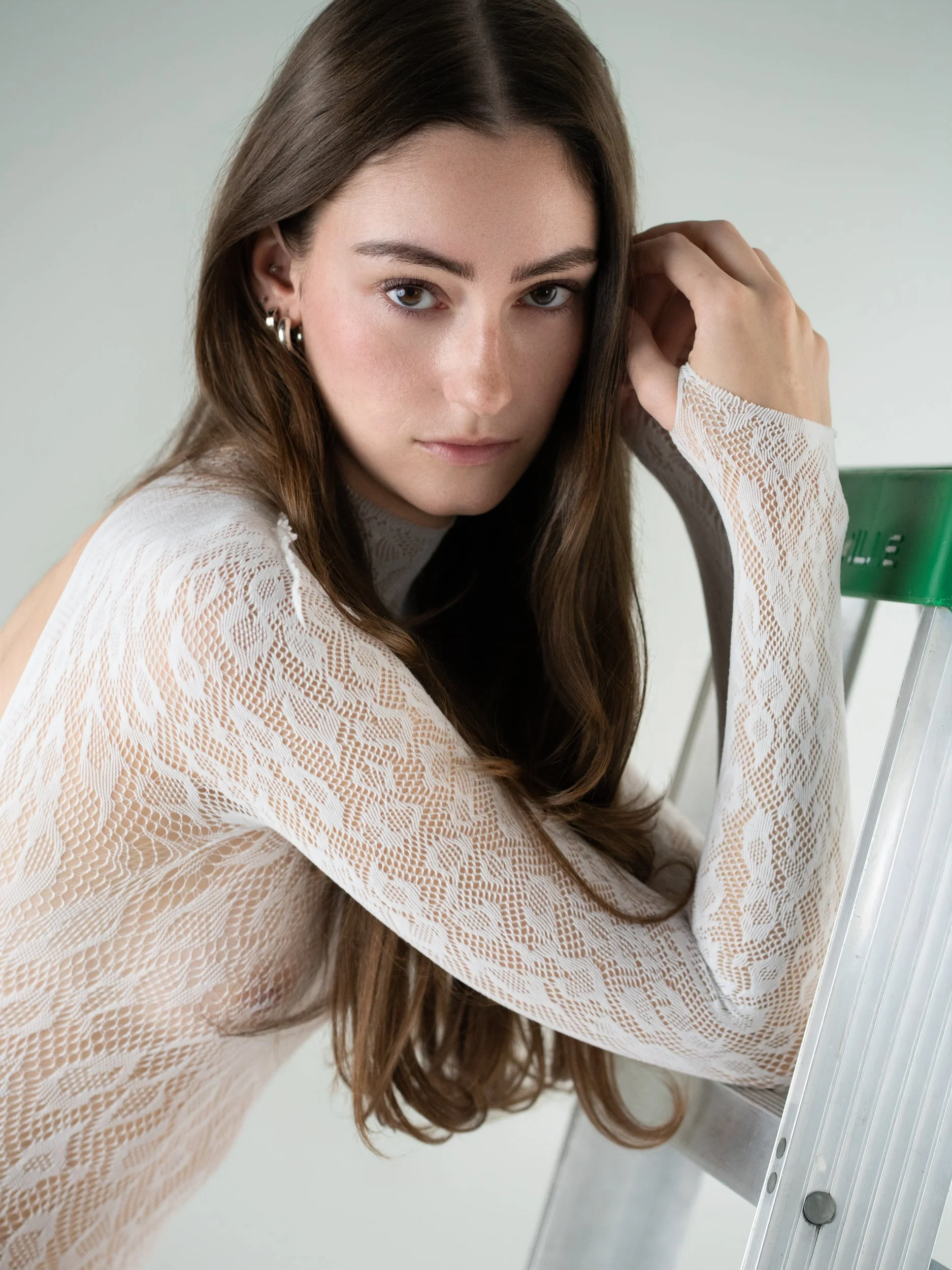 Portrait of a woman with long brown hair in a sheer white lace top, resting her head on her hand against a stepladder.