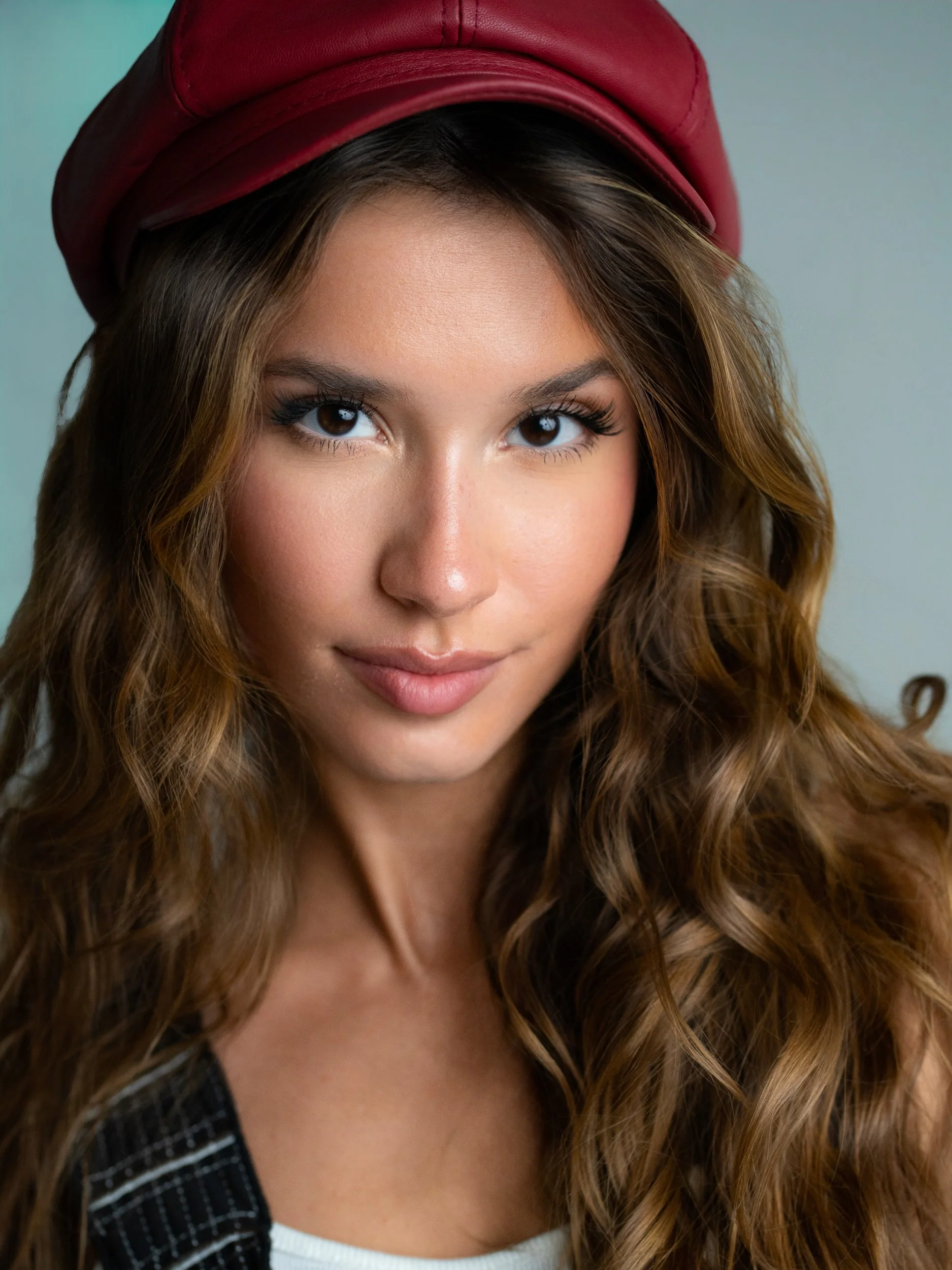 Bryanna — direct gaze, slight smile, deep-red leather baker boy cap over wavy honey-brown hair, self-styled, soft studio light.