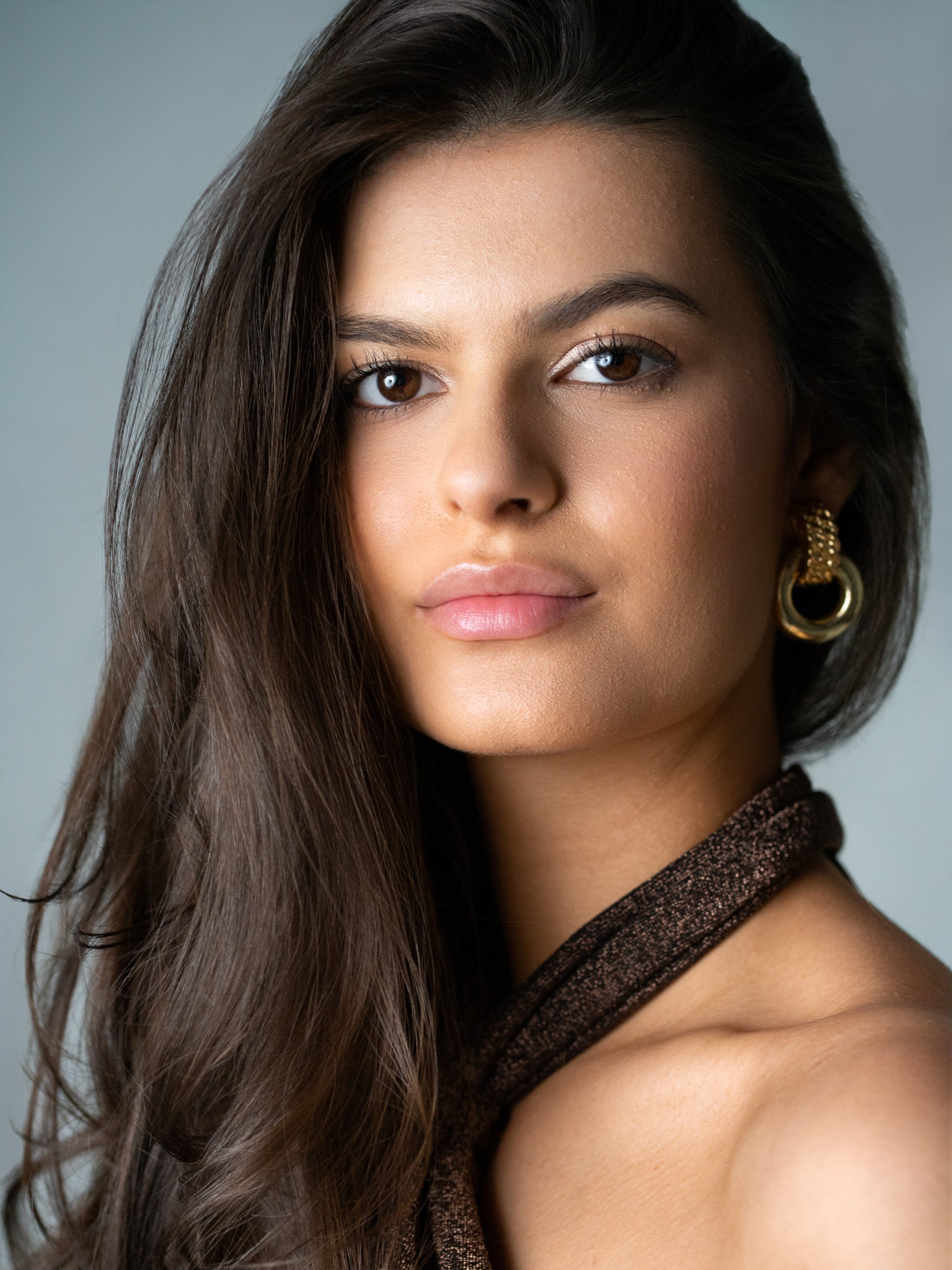 Beauty portrait of a brunette model in a bronze halter top and gold hoop earring against a grey backdrop, direct gaze to camera.