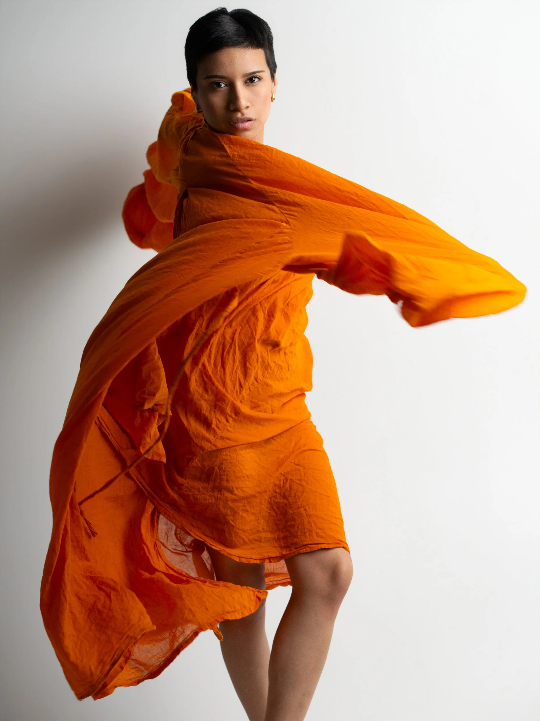 Amanda, portrait session at 9 Studios, SoHo NYC — full-length editorial, orange linen fabric in motion wrapping the body, glance back over shoulder, white background, May 2026