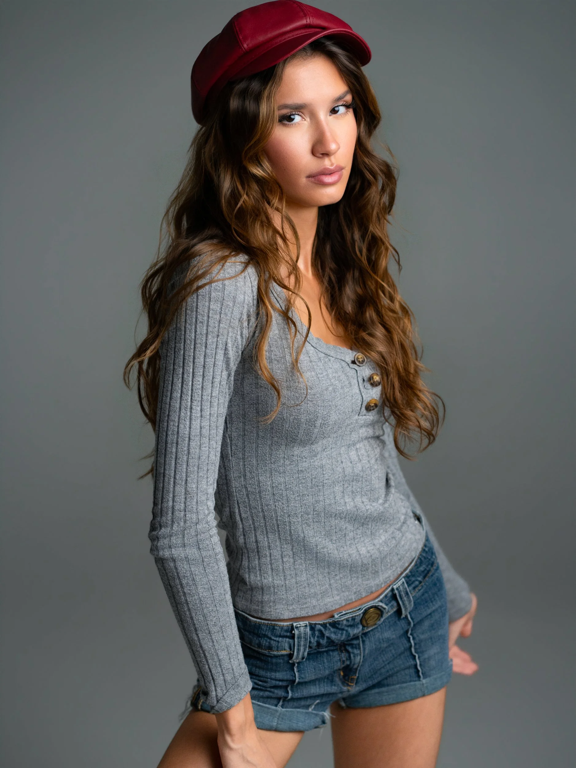 Bryanna — standing three-quarter turn, gaze to camera, red baker boy cap, grey henley, denim shorts, grey seamless.