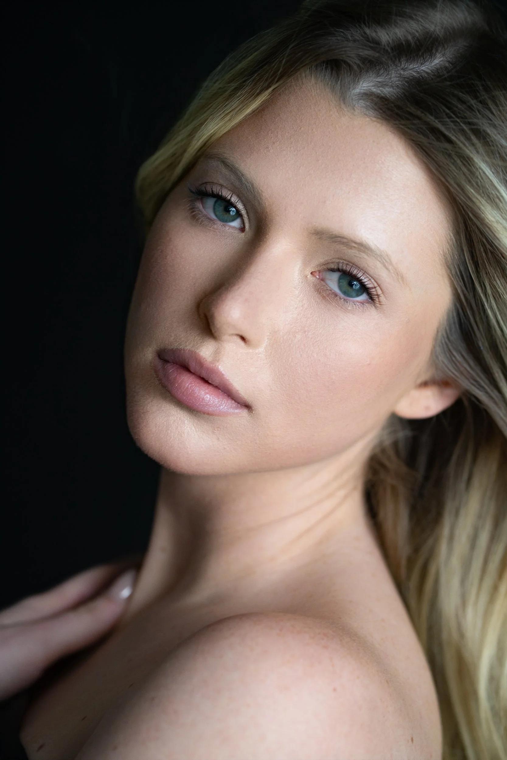Ava with a soft, level gaze and luminous skin, green eyes and tousled blonde hair, hand at her shoulder, low-key dark studio.