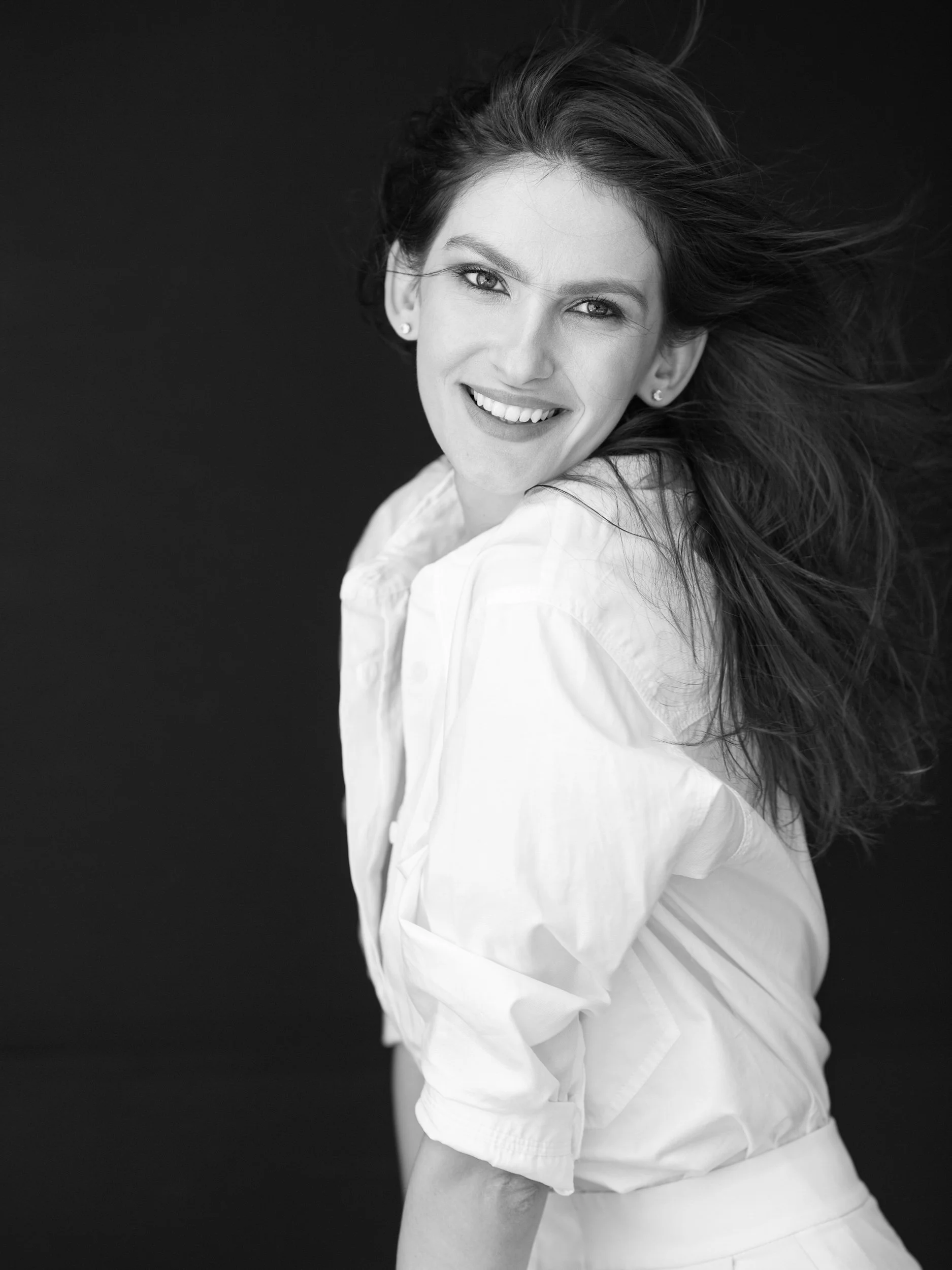 Olga, black and white portrait mid-turn with joyful smile, windswept dark hair, white shirt, dark studio background