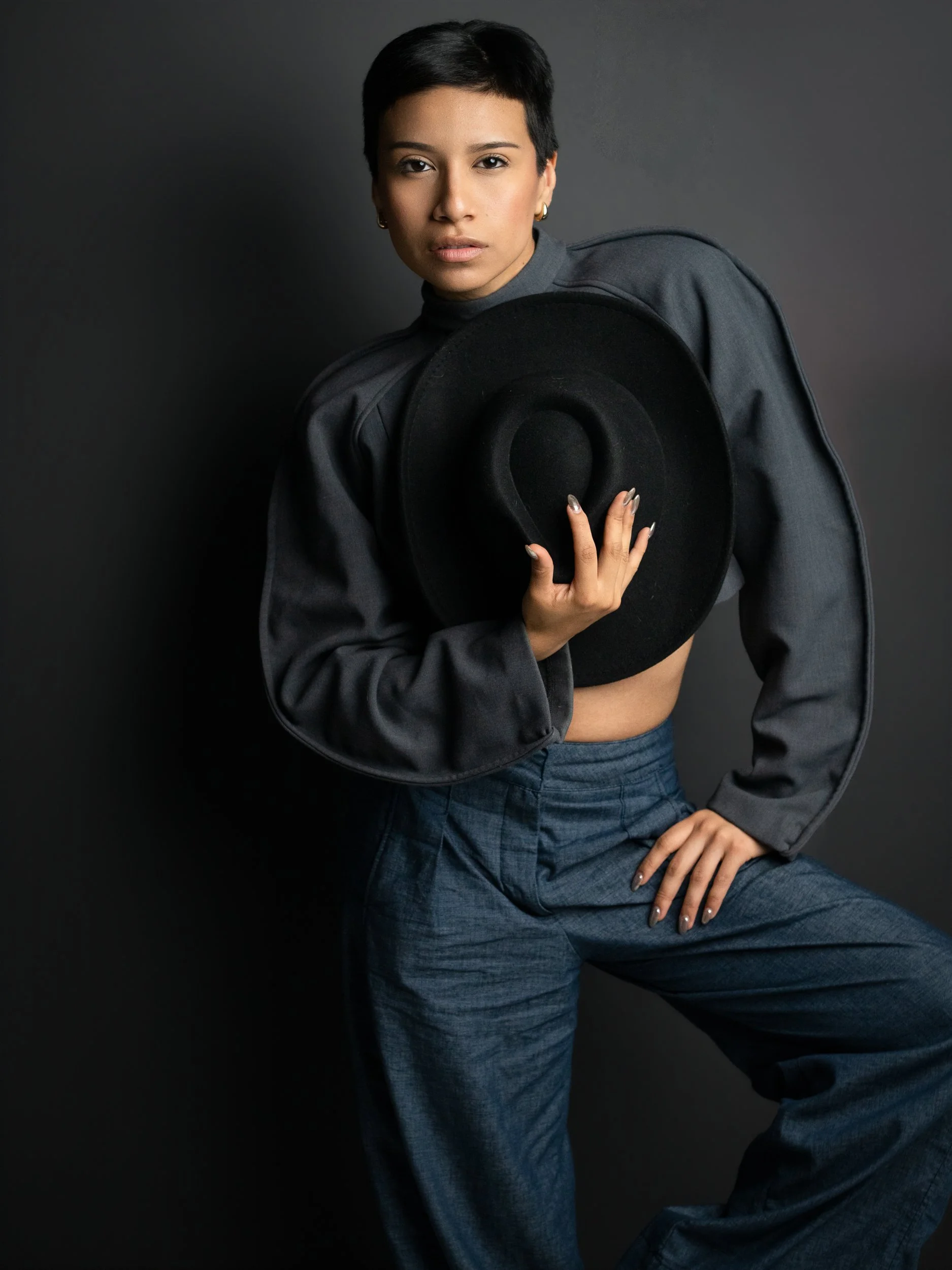 Amanda, portrait session at 9 Studios, SoHo NYC — three-quarter editorial, structured crop jacket, wide-leg trousers, wide-brim black hat held at chest, chrome nails, dark gray background, May 2026
