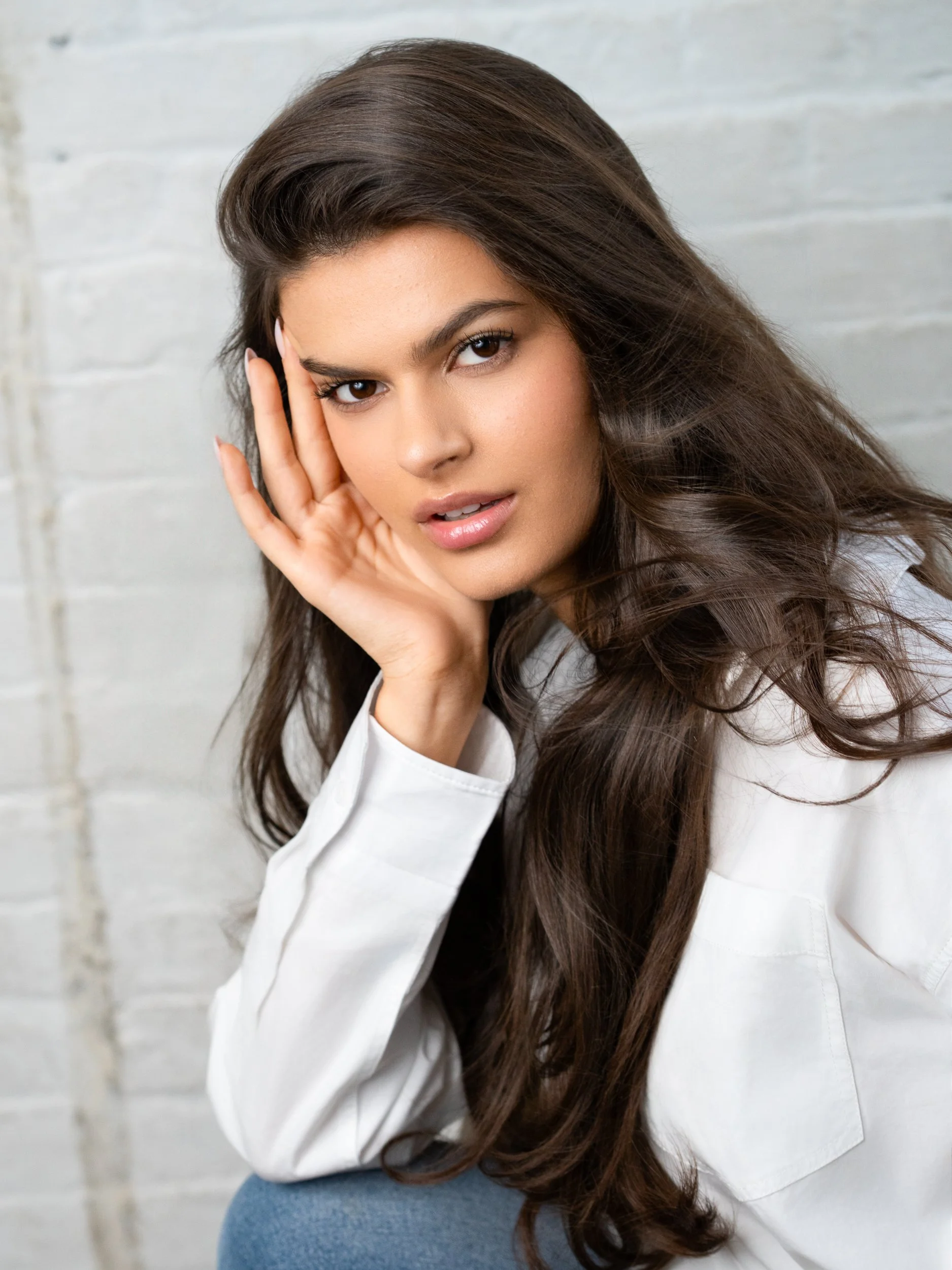 Close beauty portrait of a brunette model in a white shirt against a whitewashed brick wall, hand framing her face, gaze to camera.
