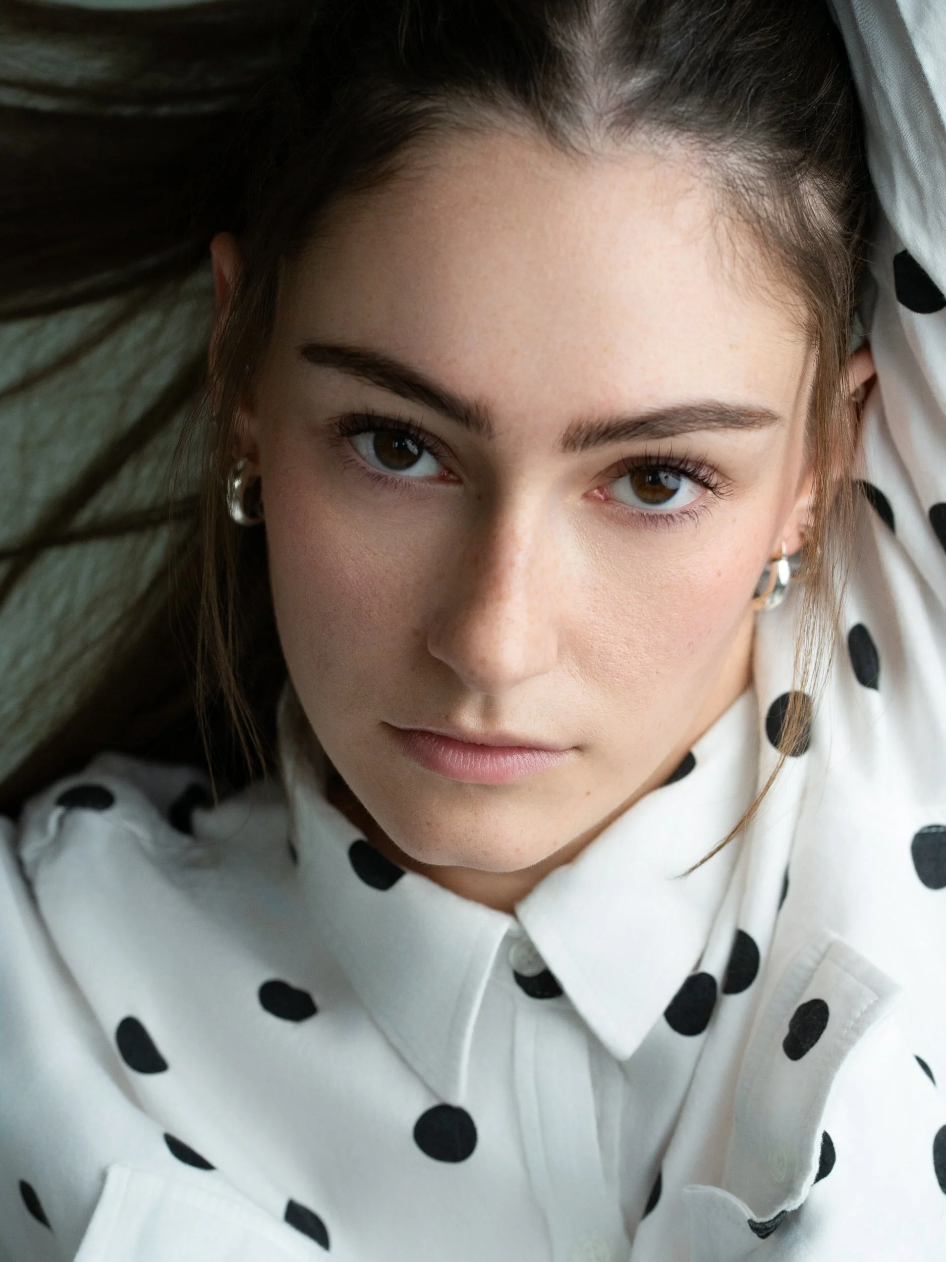 Monica in an overhead close-up, steady direct gaze, freckled skin and silver hoop earrings, black-and-white polka-dot shirt, dim studio.