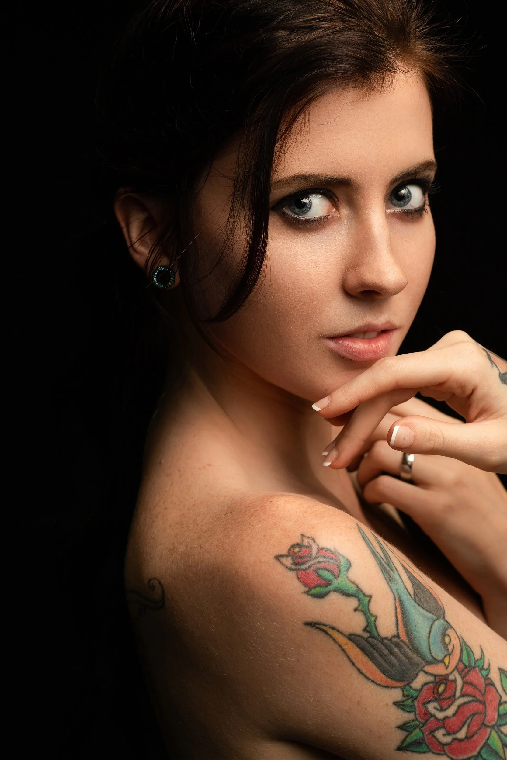 Effy turned away with a sharp sideways gaze, brunette hair and traditional-style rose and swallow tattoo on her shoulder, low-key black studio.